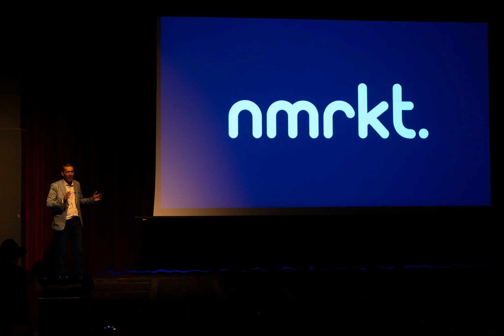 A man wearing a gray blazer and glasses standing on stage and giving a presentation. A large screen behind him displays the word 'nmrkt.' in white lowercase letters on a blue background.