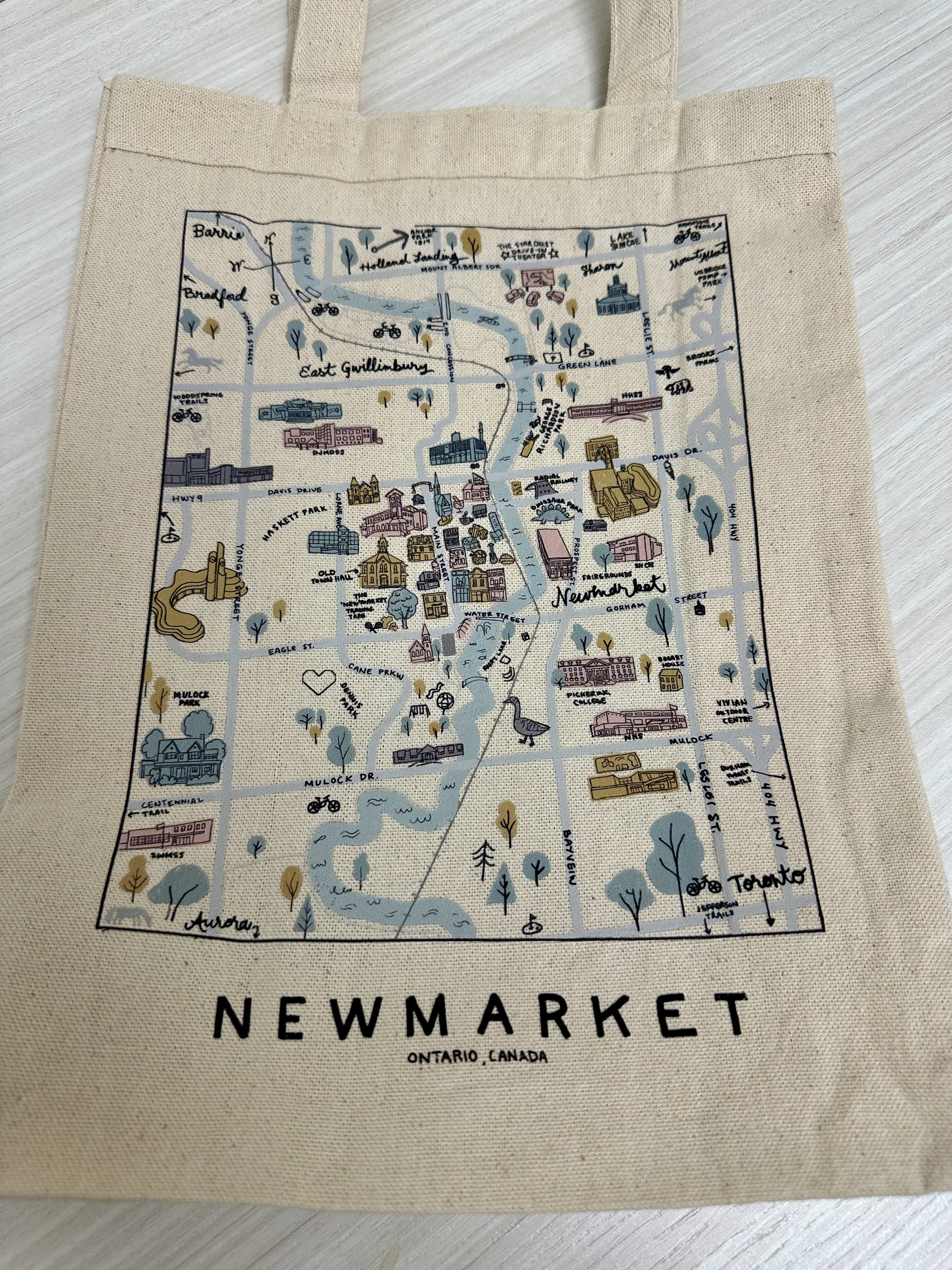 A tote bag with a illustrated map of Newmarket, Ontario, Canada, showing streets, parks, buildings, and landmarks in a stylized layout.