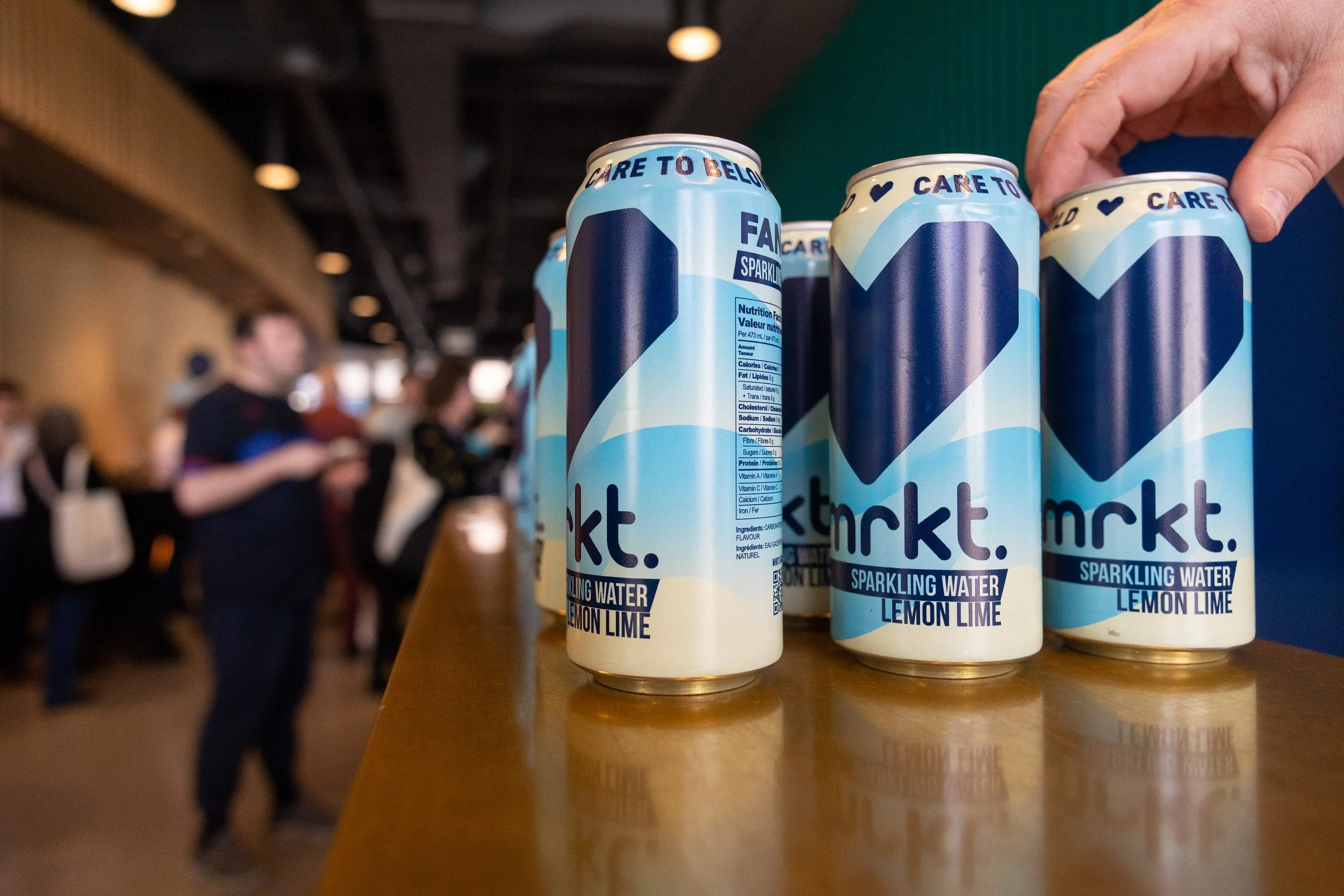 Cans of nmrkt. sparkling water lemon lime on a wooden counter, with an out-of-focus background of people in a bustling event setting.