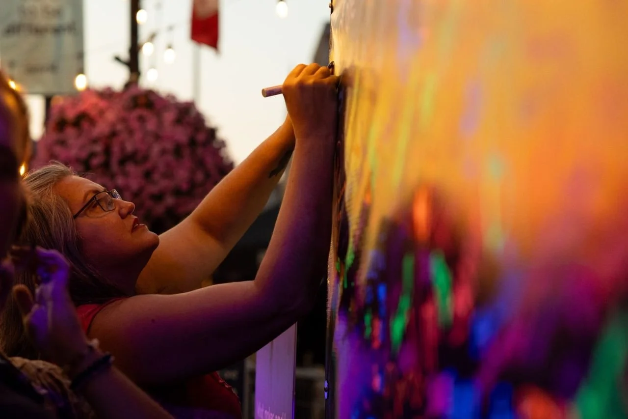 A woman with glasses writing or drawing on a vibrant, colorful wall or board during an outdoor event in the evening.