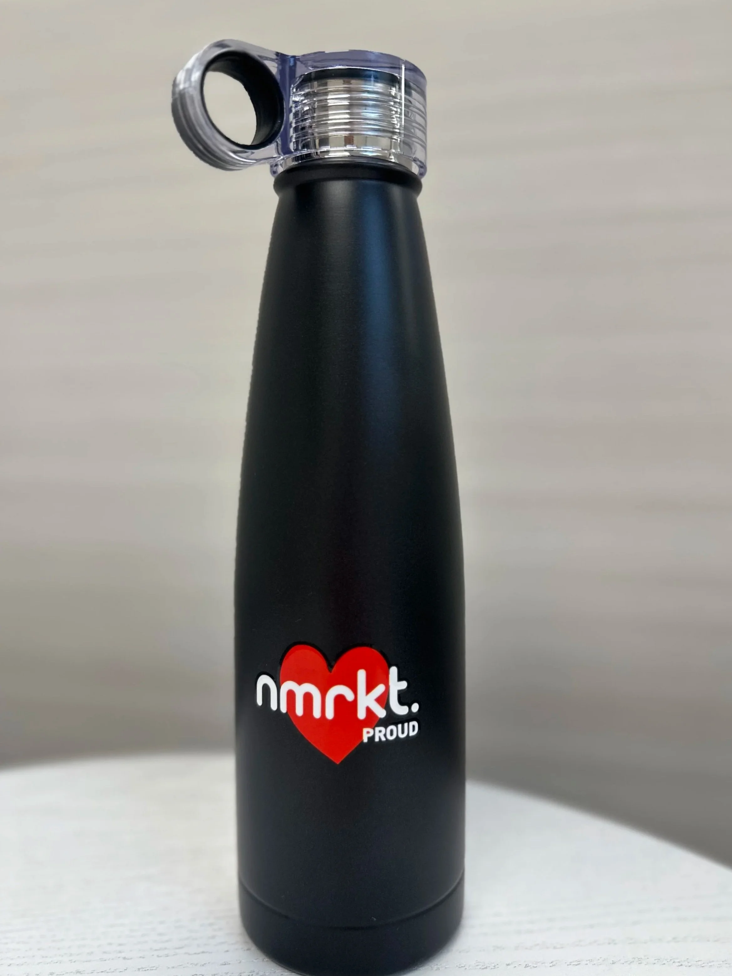 Black water bottle with a metal cap and a black loop, featuring a red heart graphic and the words 'nmrkt. PROUD' printed on it.