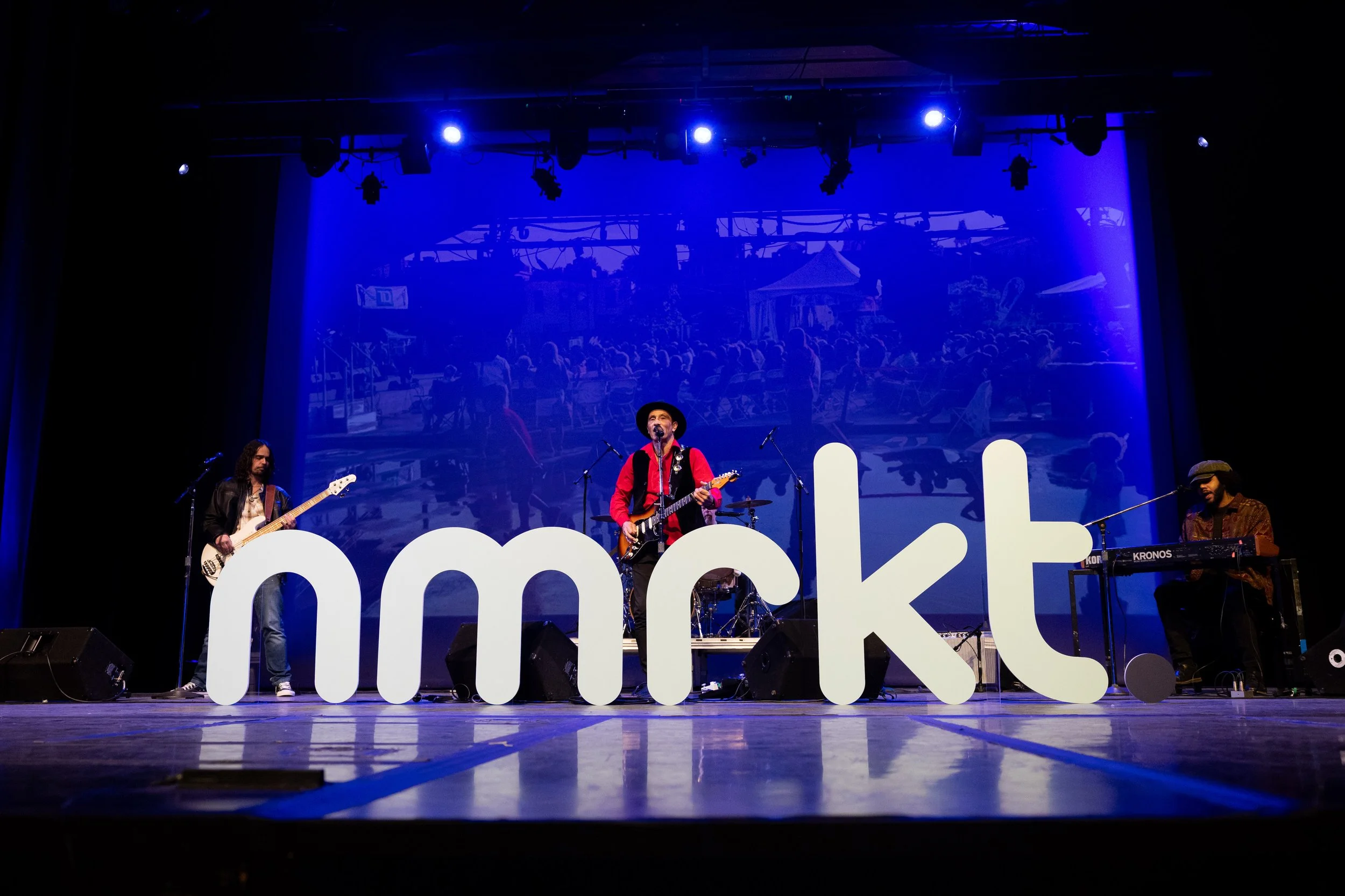 Musicians performing on stage at a concert, with a large 'nmrkt.' sign in front, a blue background projection of an outdoor scene, and stage lighting.