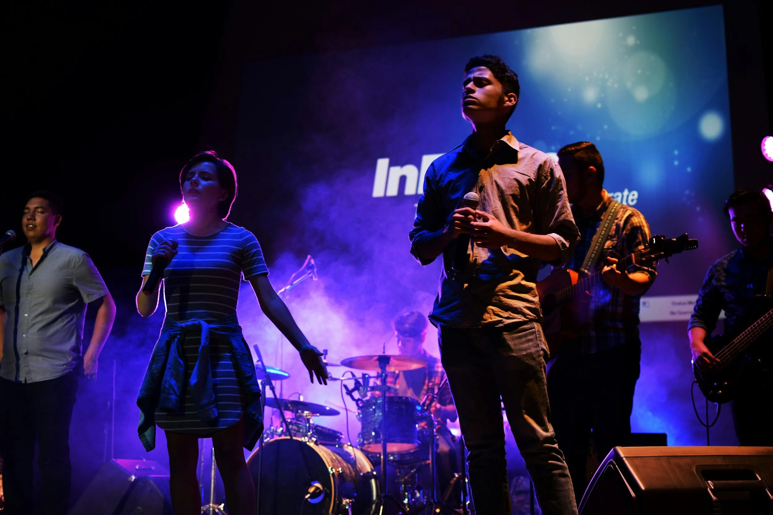 A group of young performers on a dimly lit stage with blue and purple lighting, some singing into microphones and others playing instruments, with a large screen in the background displaying the word 'Inc'.