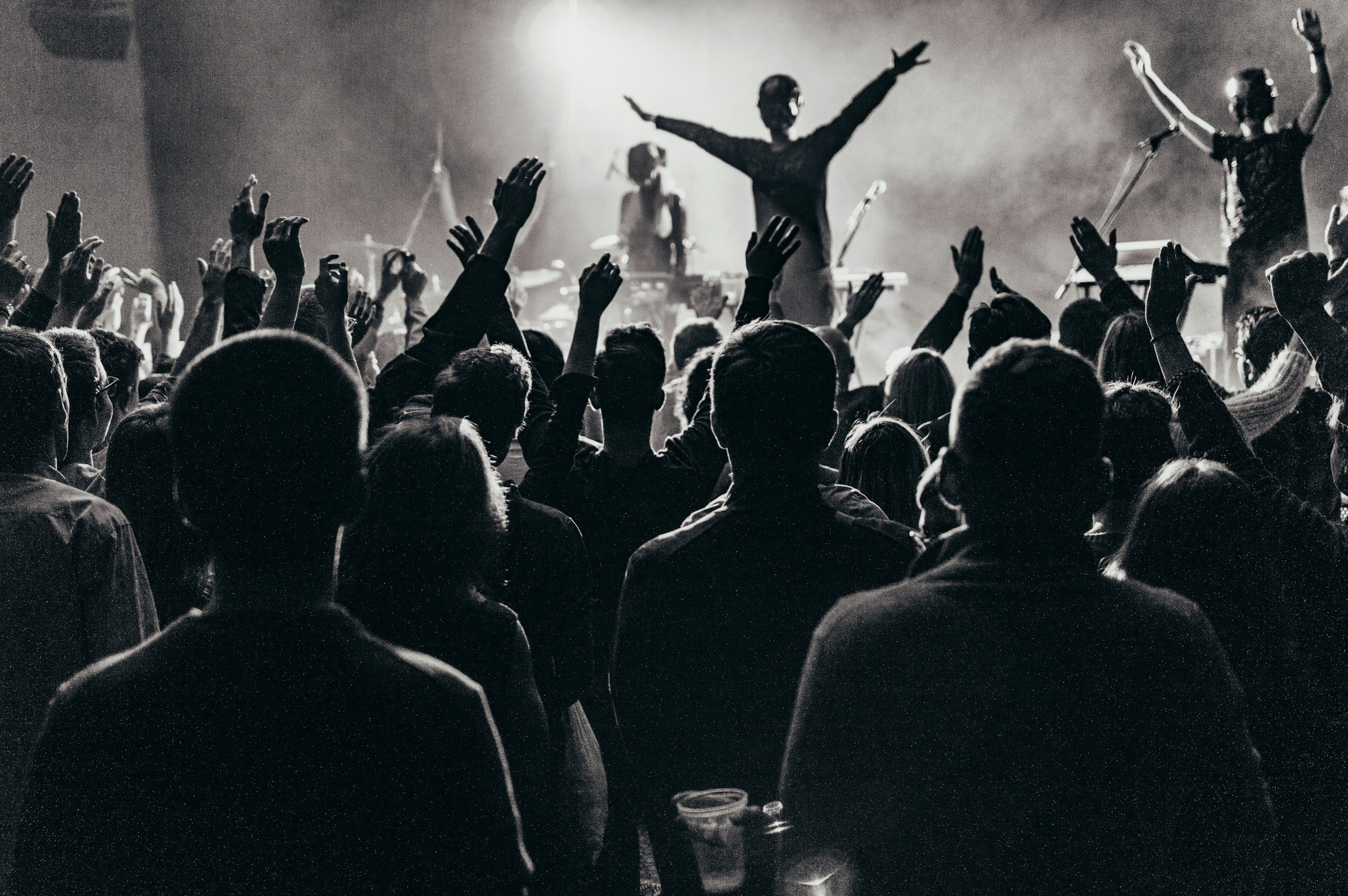 A crowded black-and-white photo of a concert or performance, with people raising their hands in the audience and performers on stage.