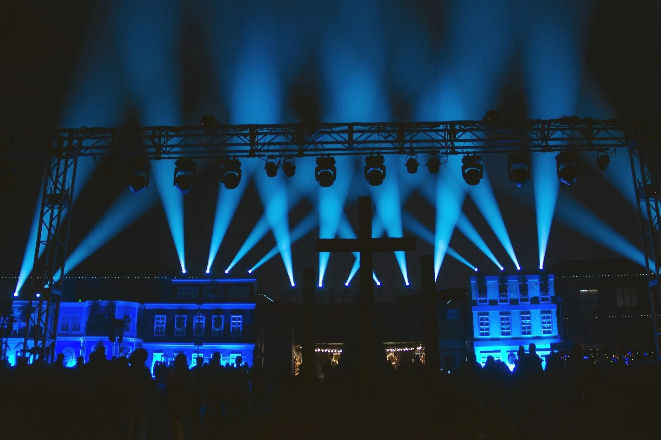 Night scene of a church stage illuminated with blue lights and spotlights, with a cross in front and a crowd of people watching.