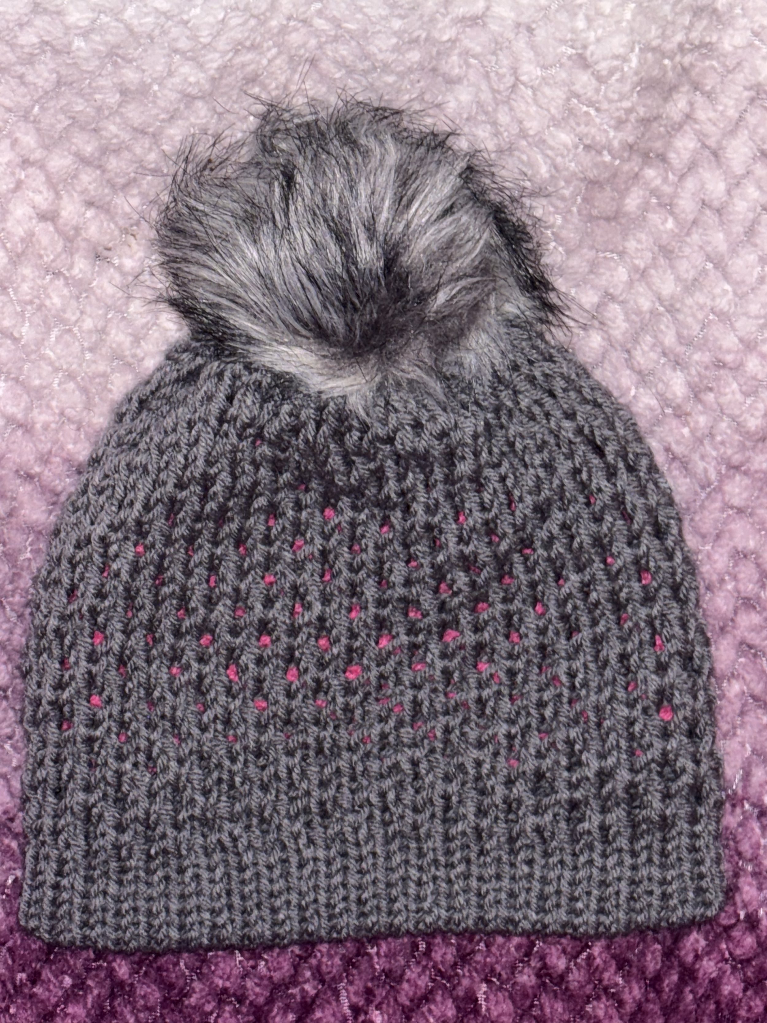 Gray knit beanie hat with a faux fur pom-pom on top, resting on a pink textured surface.