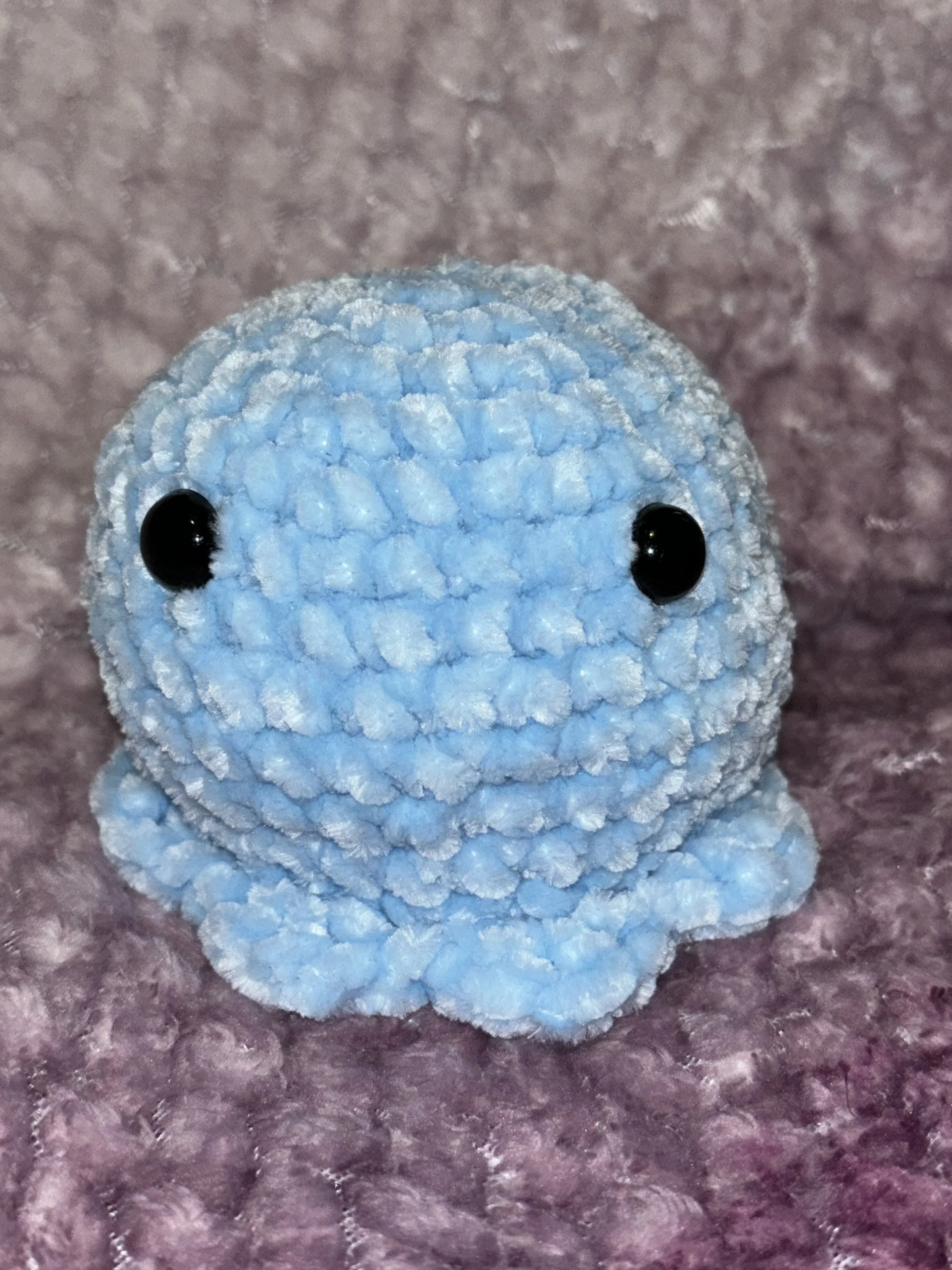 A small blue stuffed animal resembling an octopus with black eyes, sitting on a textured purple surface.