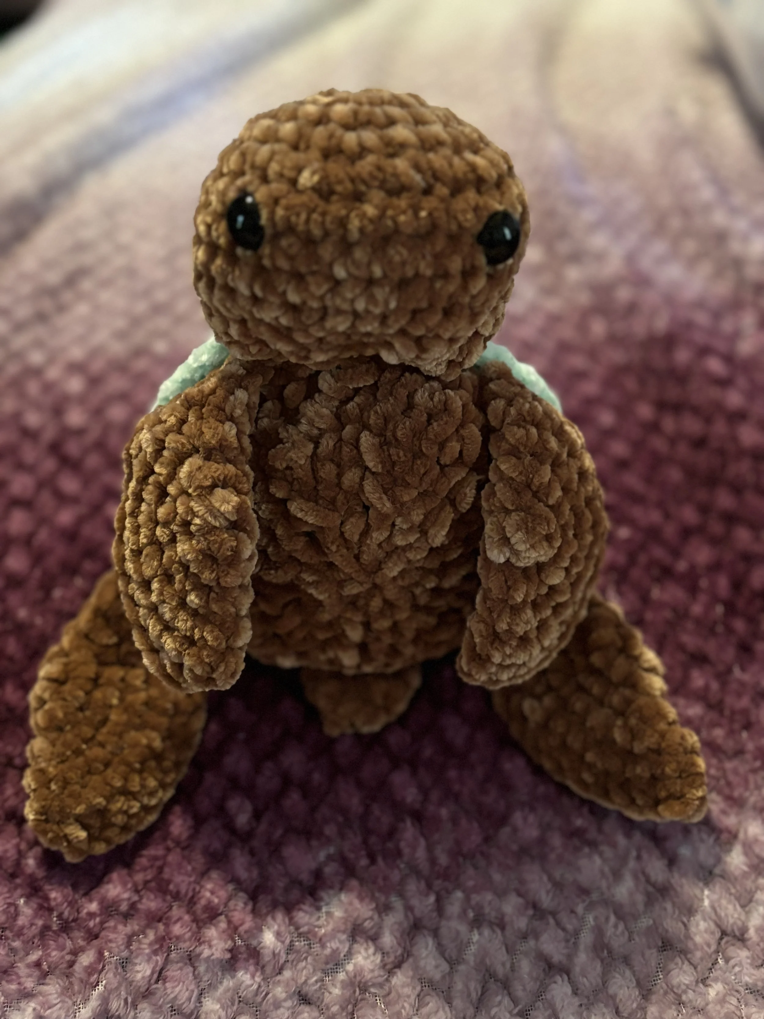 A plush teddy bear made of textured brown fabric sitting on a pink and purple patterned surface.