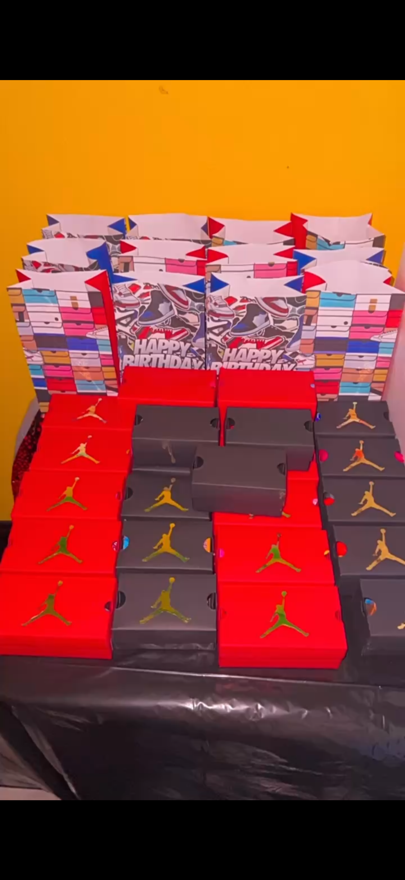 A display of multiple Nike Air Jordan sneakers boxes with a combination of red and black boxes, stacked in front of a background of colorful gift bags and a yellow wall.