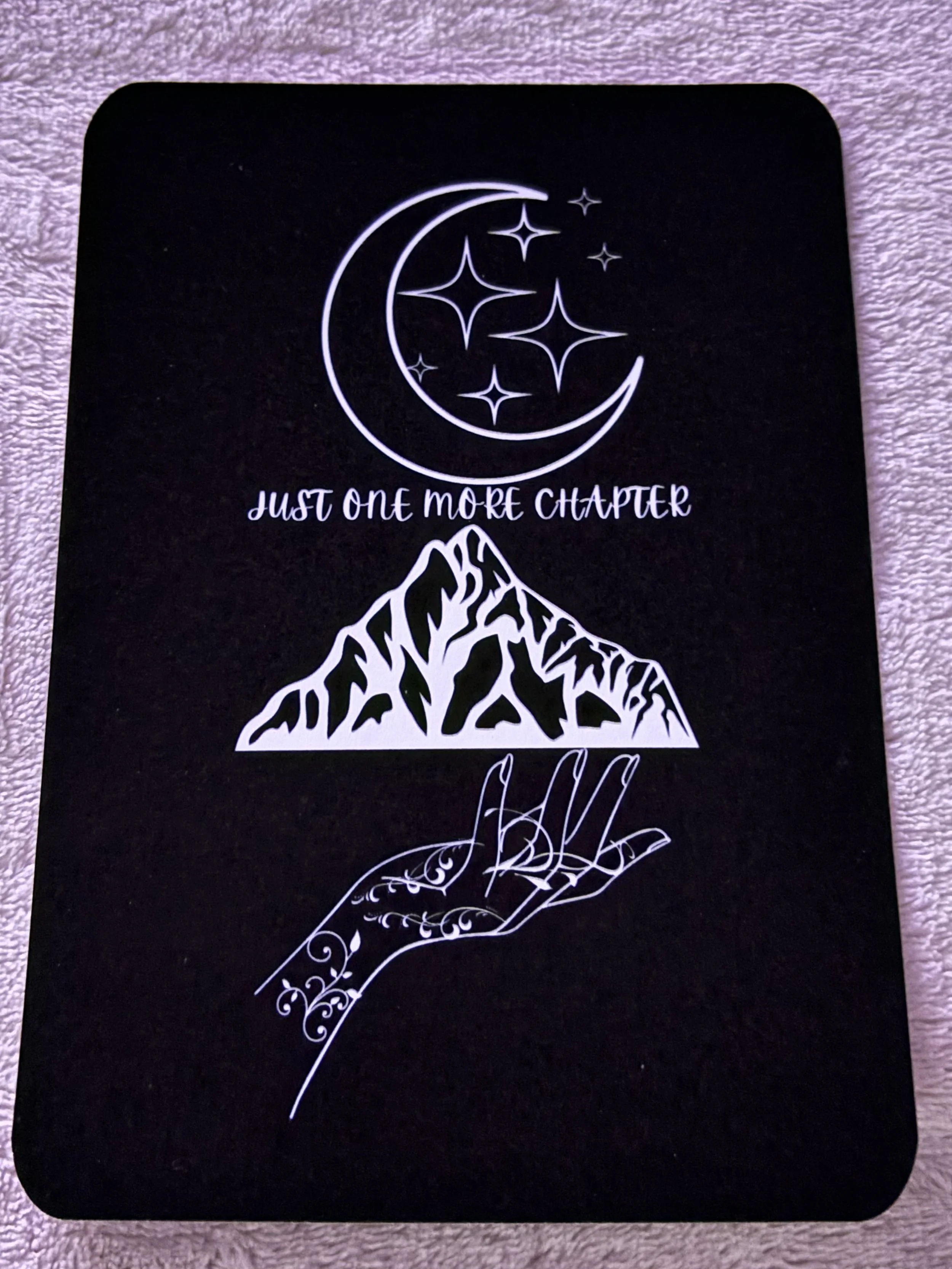 A black notebook cover with white illustrations of a crescent moon and stars, a mountain, and a decorative hand, along with the phrase "Just one more chapter".