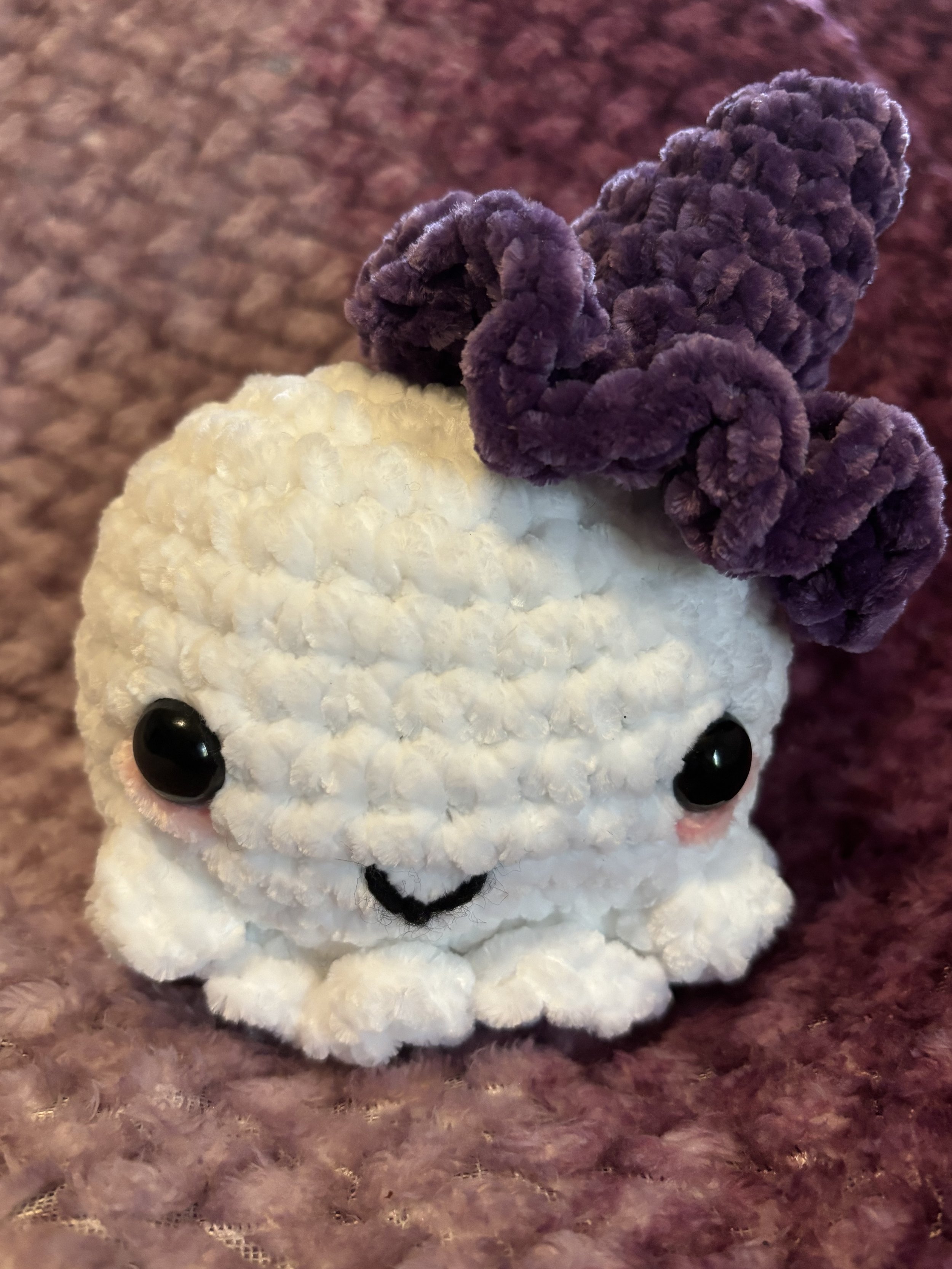 A small, plush crocheted toy resembling a cute white animal with big black eyes, a small smile, and purple curly hair on its head, sitting on a purple textured surface.