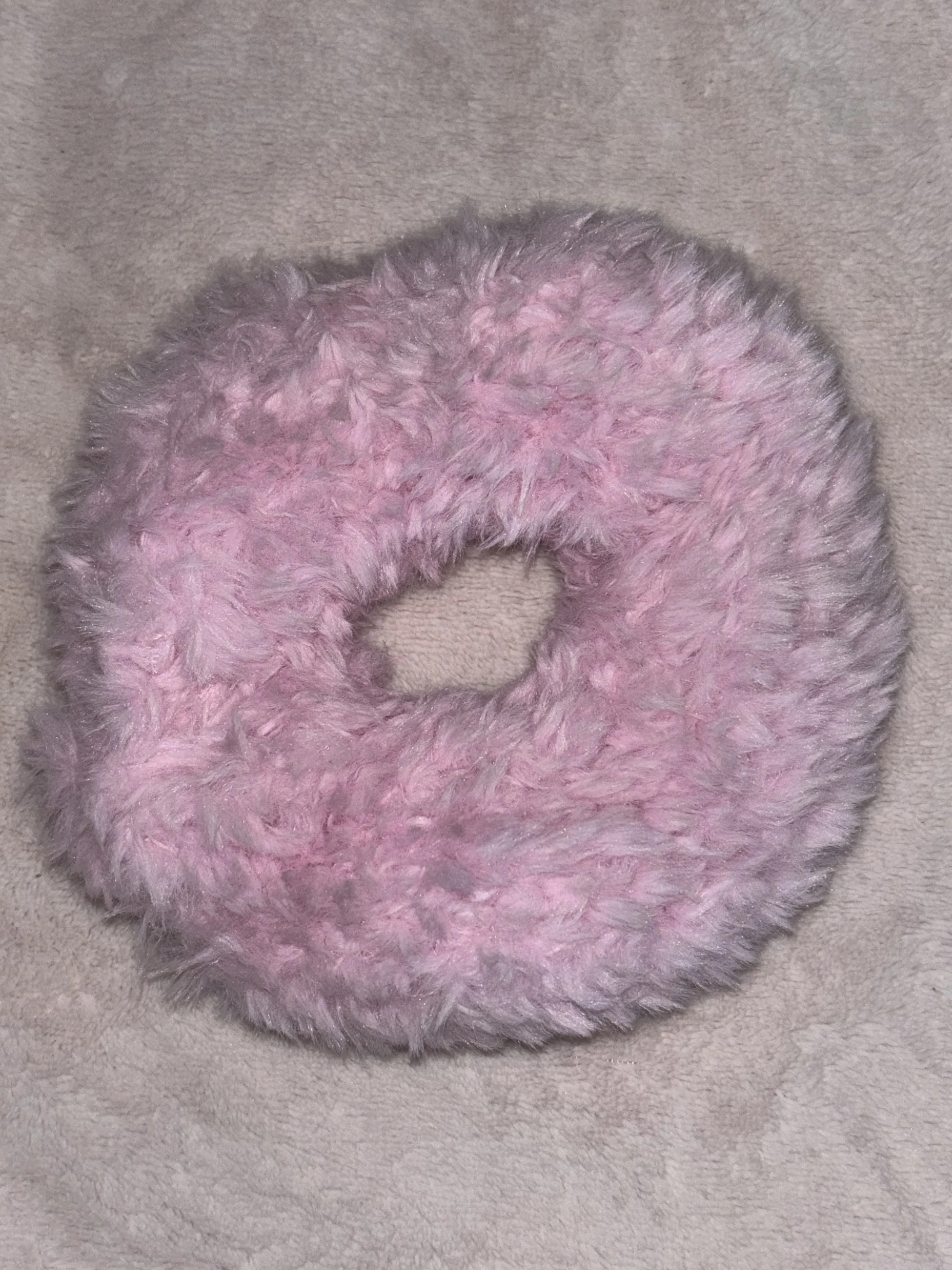 A round, fluffy pink and gray fuzzy ring, possibly a scrunchie or decorative accessory, placed on a beige surface.