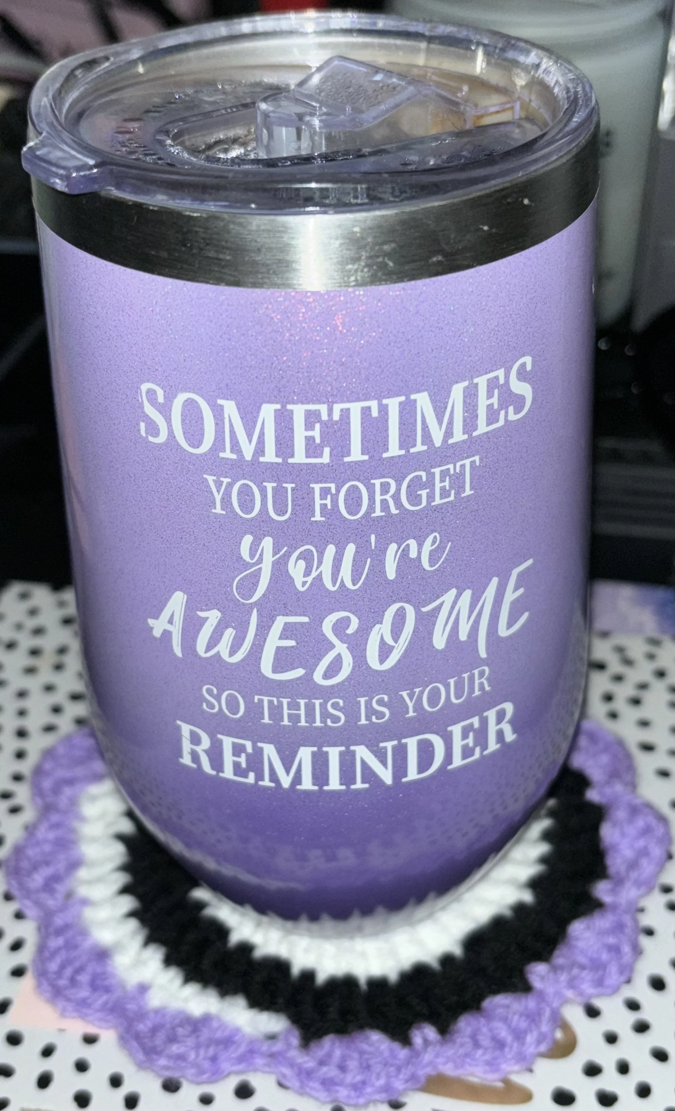 A purple insulated tumbler with a clear plastic lid, sitting on a black, white, and purple crocheted coaster, displaying a motivational quote in white text.