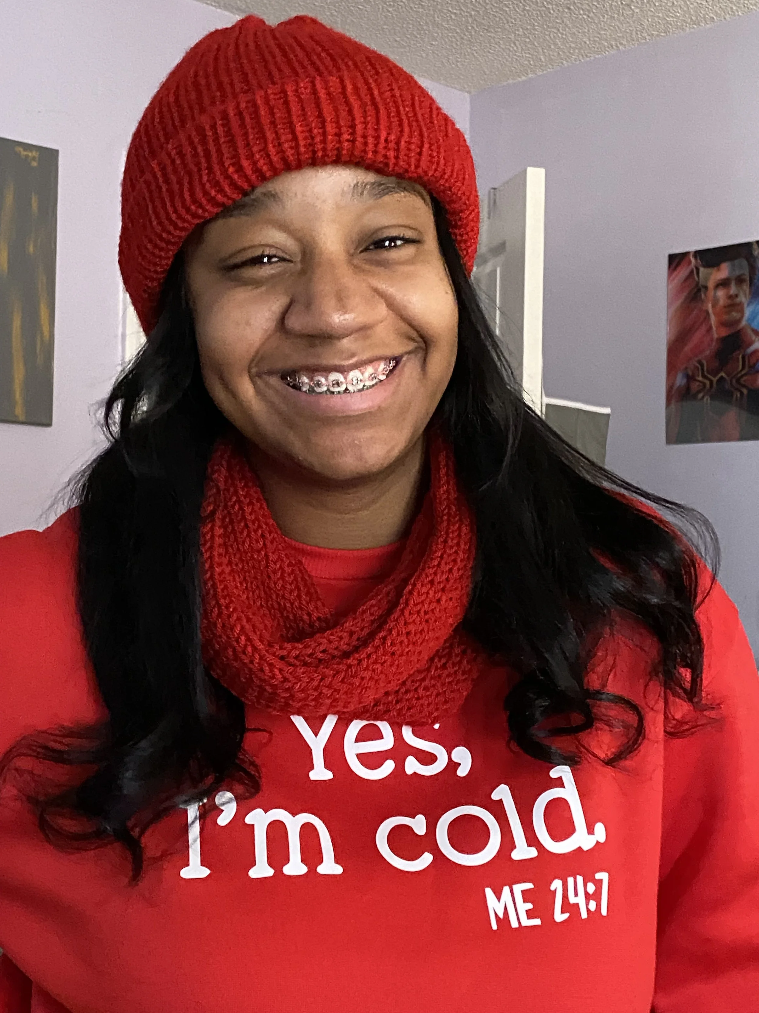 A young woman smiling, wearing a red knit beanie and matching red sweatshirt with white text that says, 'Yes, I'm cold. ME 24:7.' She has dark wavy hair and braces. The background shows a light purple wall with some artwork and an open door.