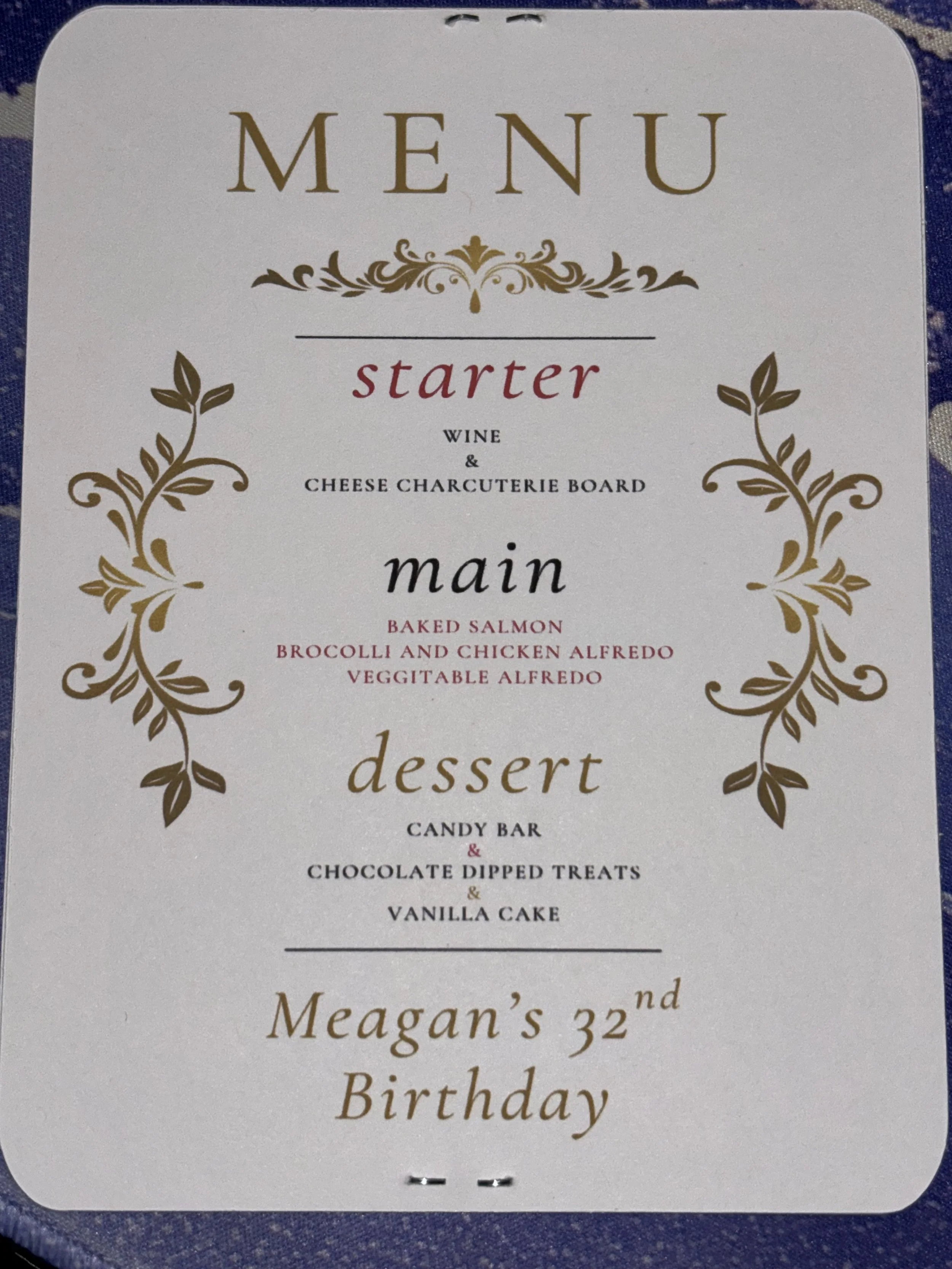 Menu card with a gold decorative border, listing starter, main, and dessert options for a 32nd birthday celebration. The starter includes wine and cheese charcuterie board; main options are baked salmon, broccoli and chicken Alfredo, and vegetable Al