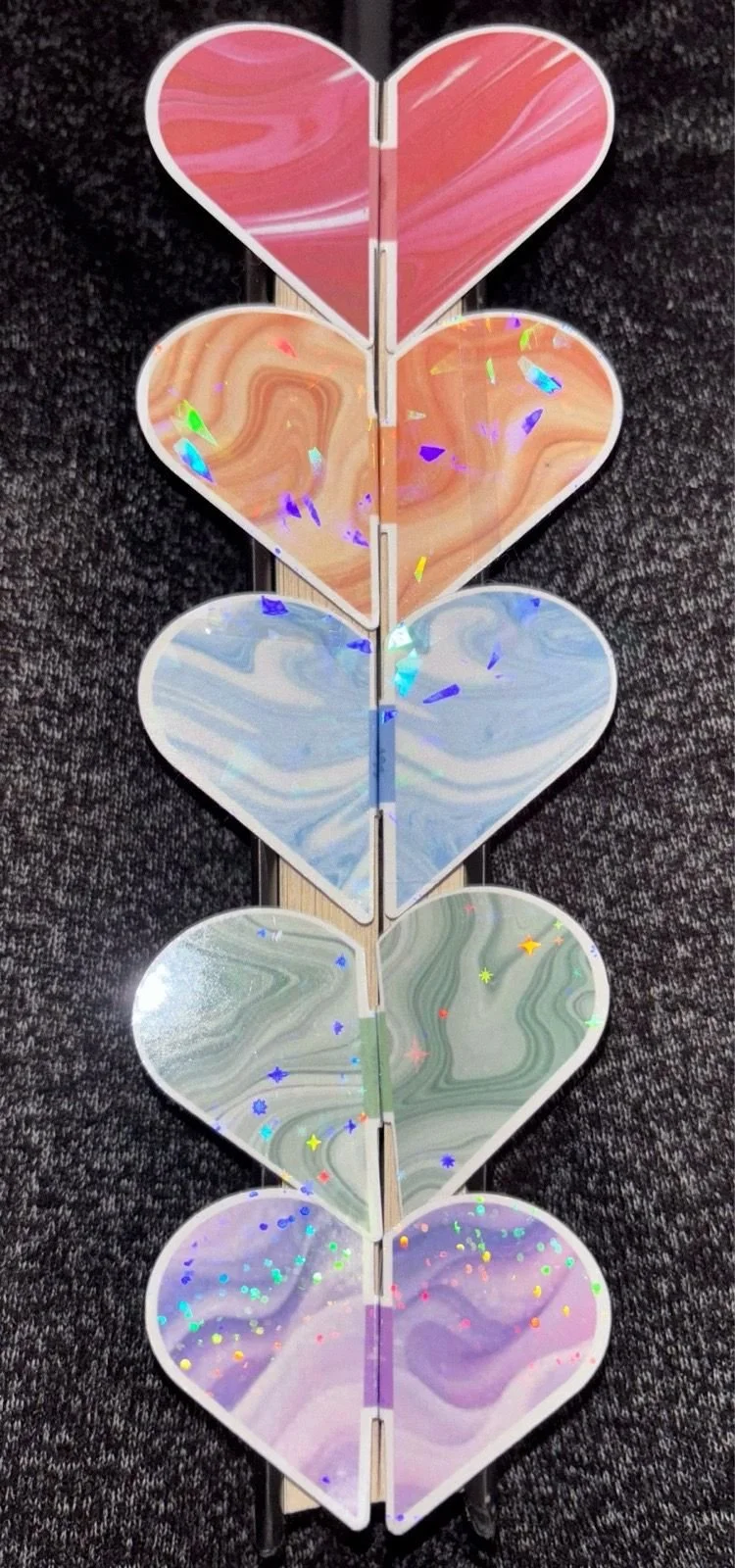 A vertical array of six paper hearts with marbled patterns and holographic confetti, arranged on a black textured surface.