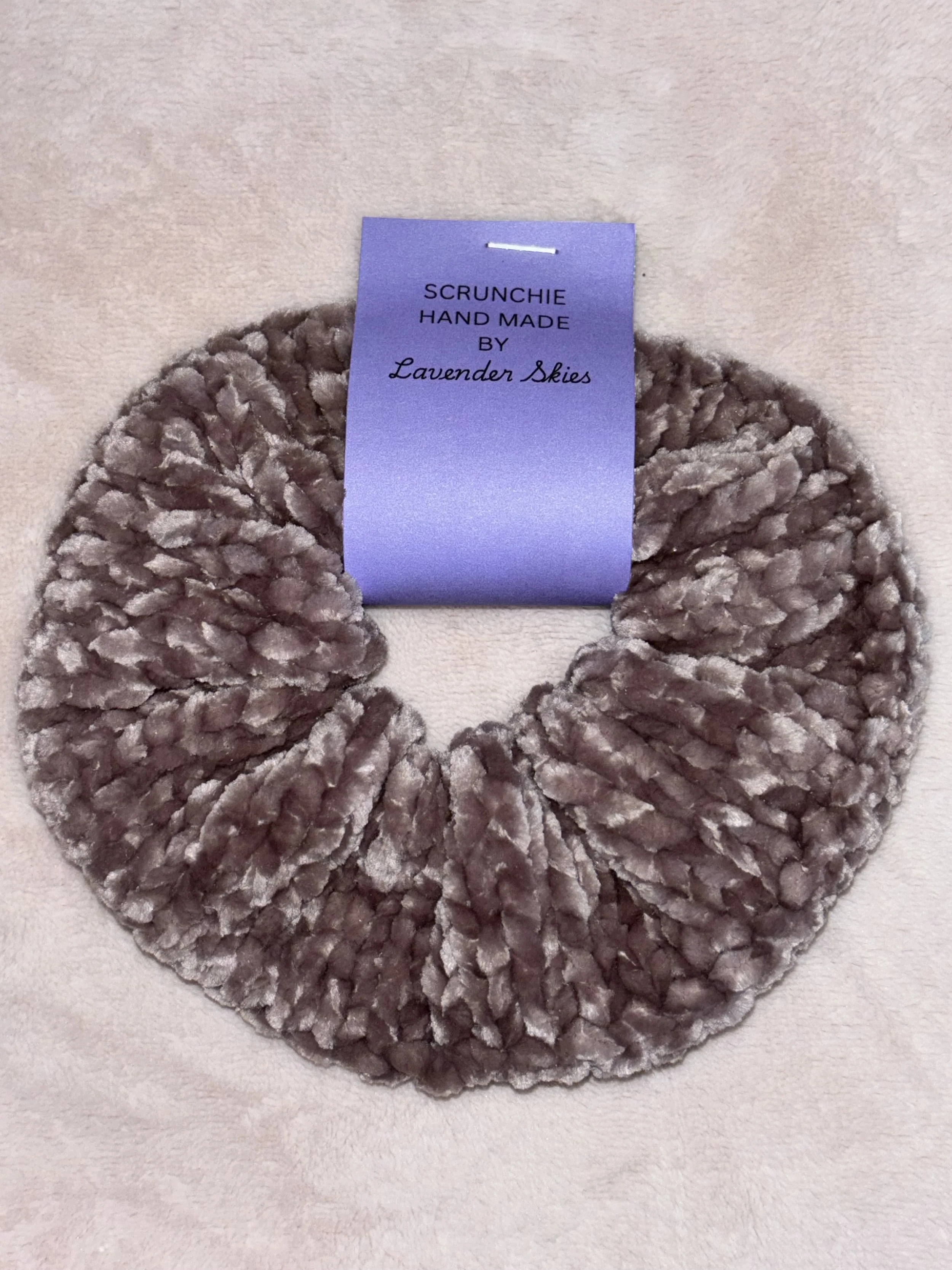 A soft, fluffy scrunchie made of plush fabric in a taupe color, with a lavender-colored tag that reads 'SCRUNCHIE HAND MADE BY Lavender Skies'.