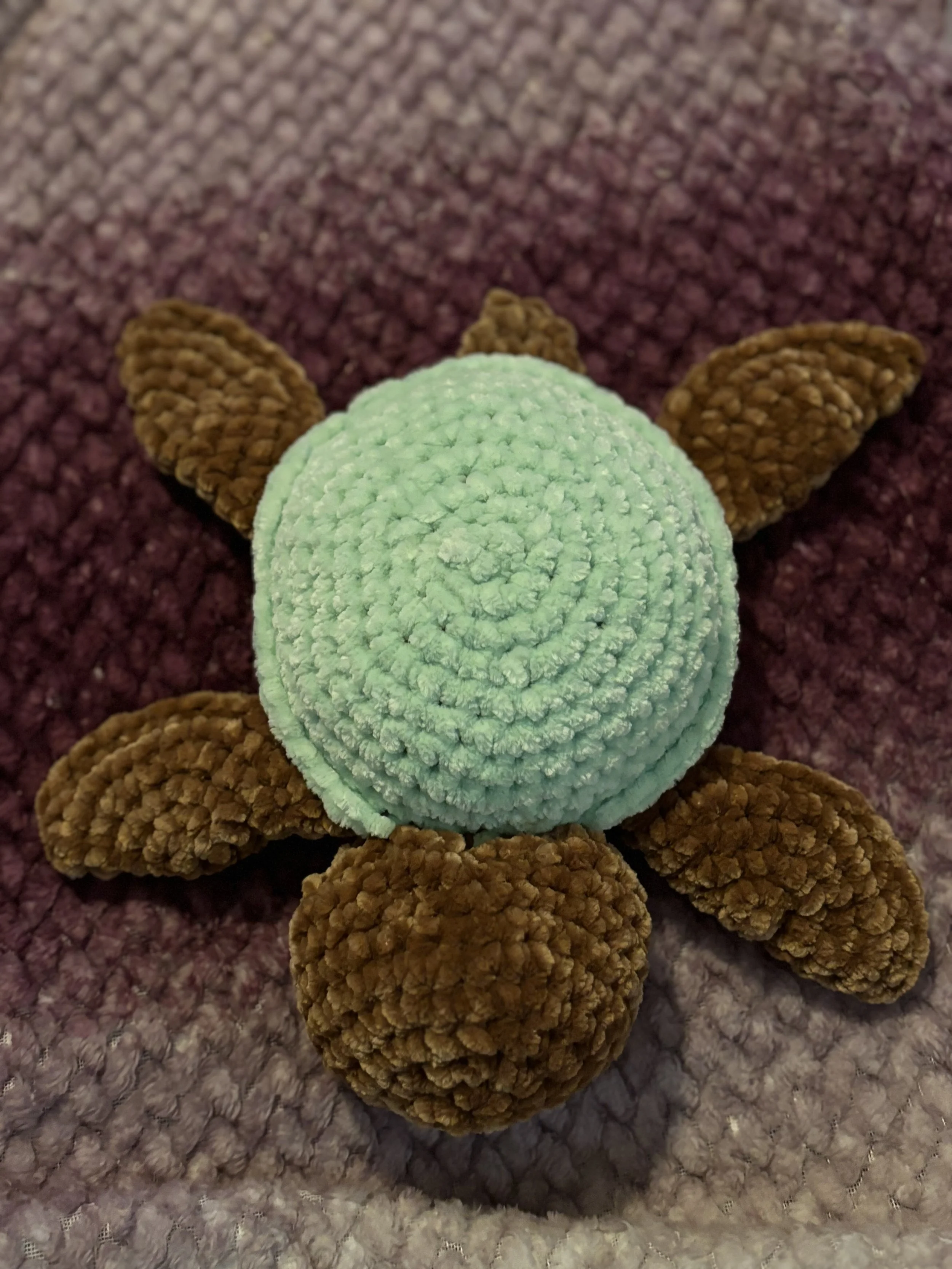 A handmade fabric turtle with a mint green shell and brown limbs, lying on a textured purple blanket.