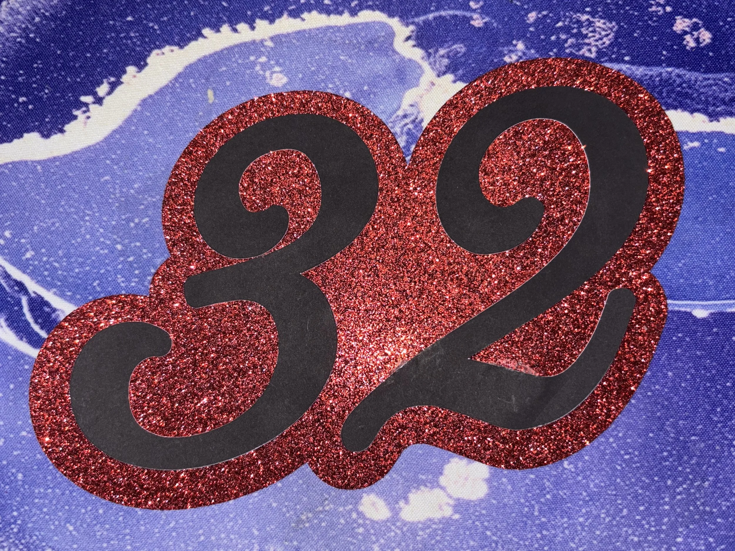 A shiny red glitter number 32 with black numbers cut out, placed against a blue and white marbled background.
