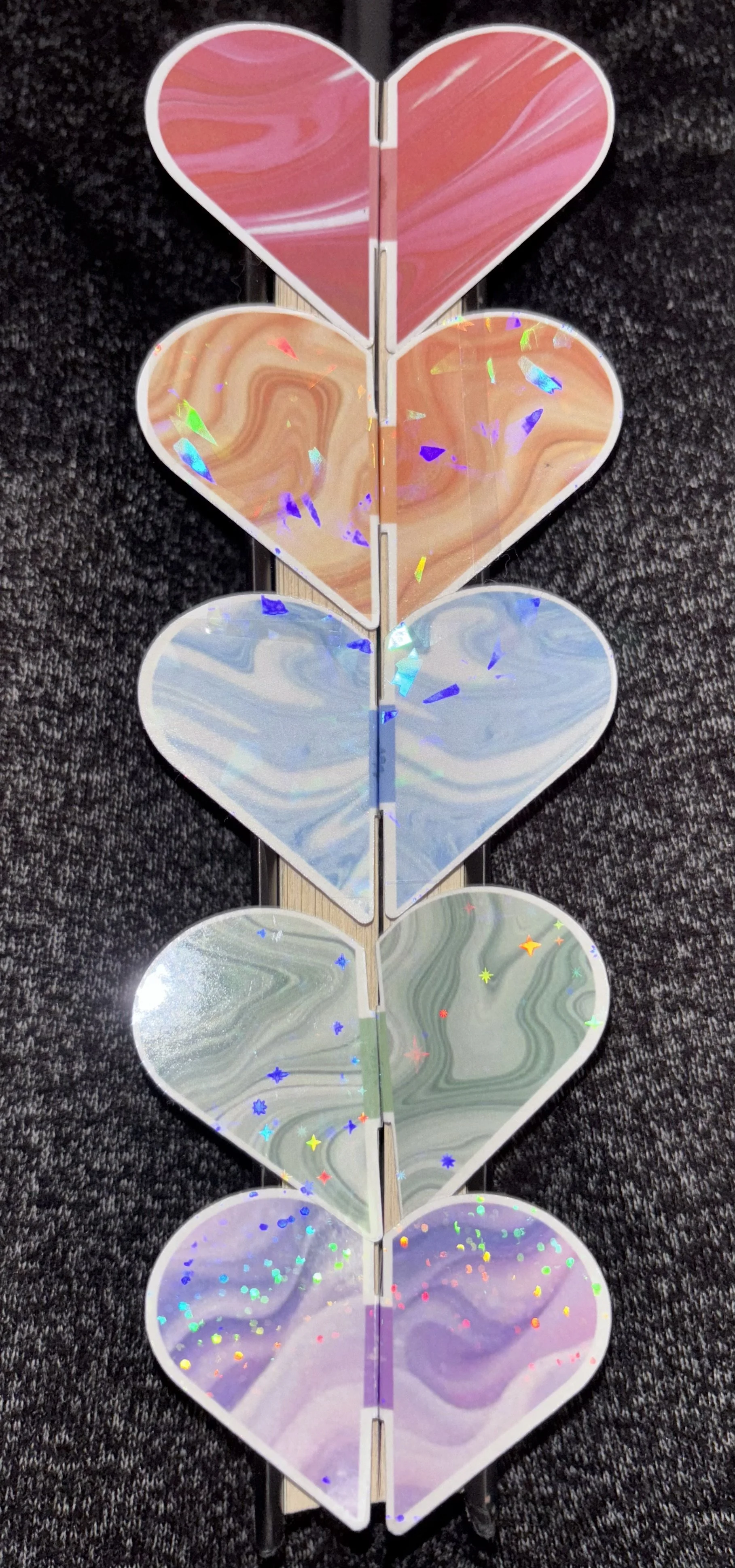 A vertical row of five iridescent heart-shaped mirrors with holographic confetti, resting on a dark textured surface.