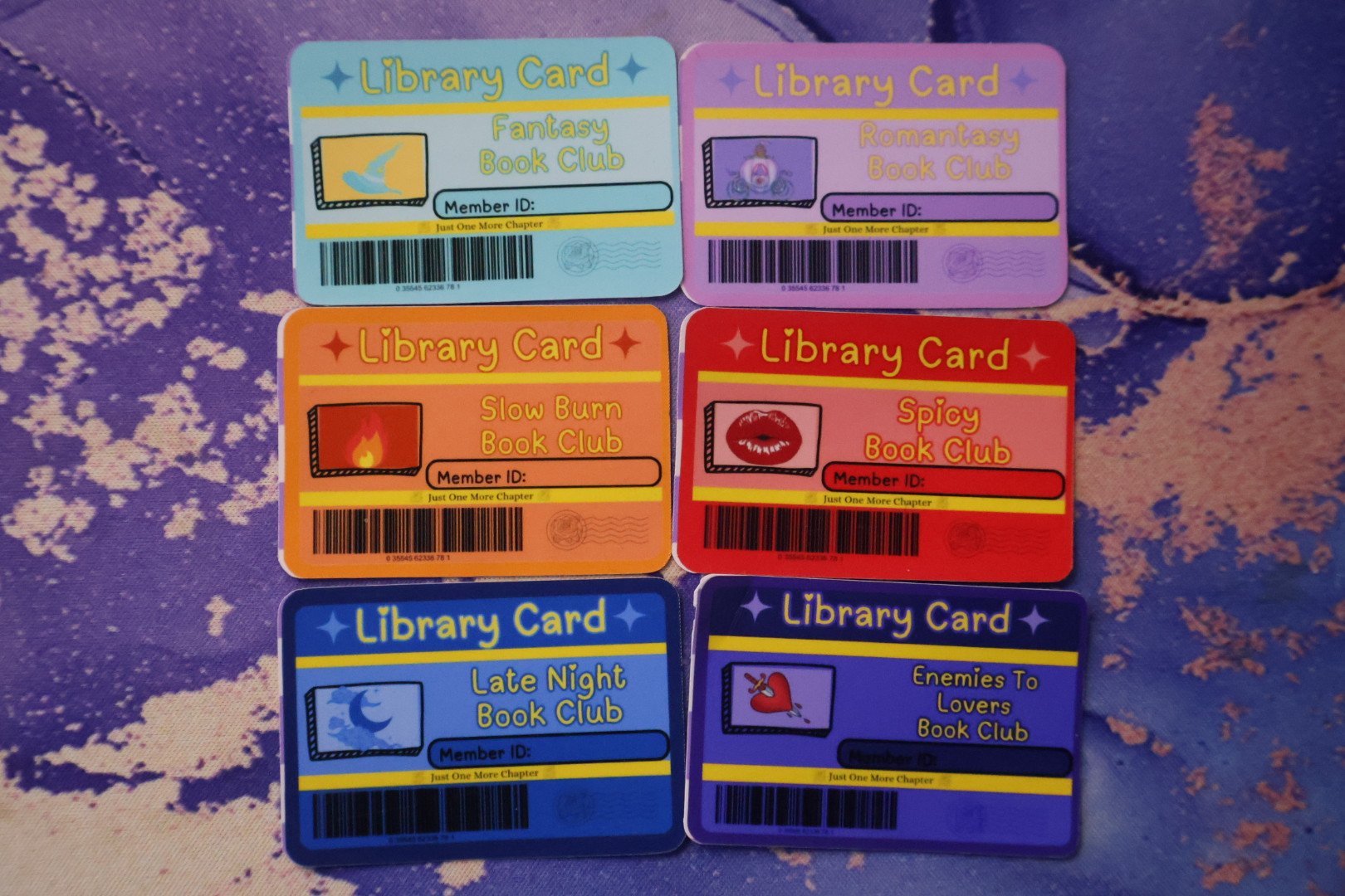 Five colorful library cards, each with a unique theme and illustration, arranged on a purple and pink textured background. The cards are for fantasy, romance, slow burn, spicy, late night, and enemies-to-lovers book clubs.