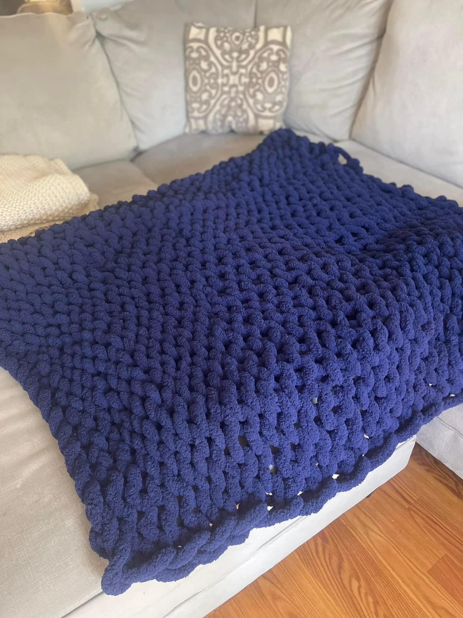 A dark blue chunky knit blanket on a light-colored sofa with pillows in the background.