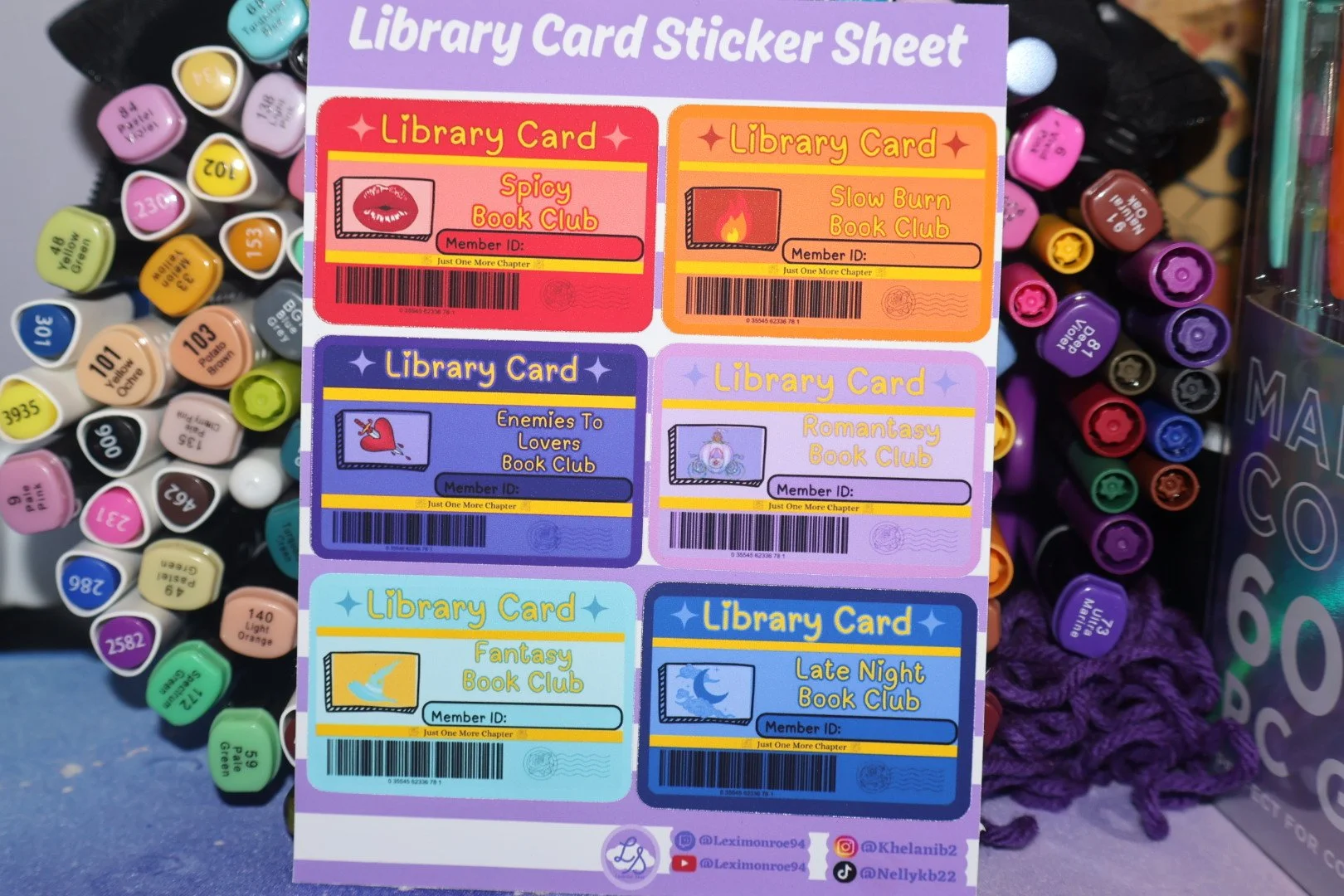 A sheet of library card stickers with six different designs featuring various book club themes, surrounded by colorful markers and other stationery.