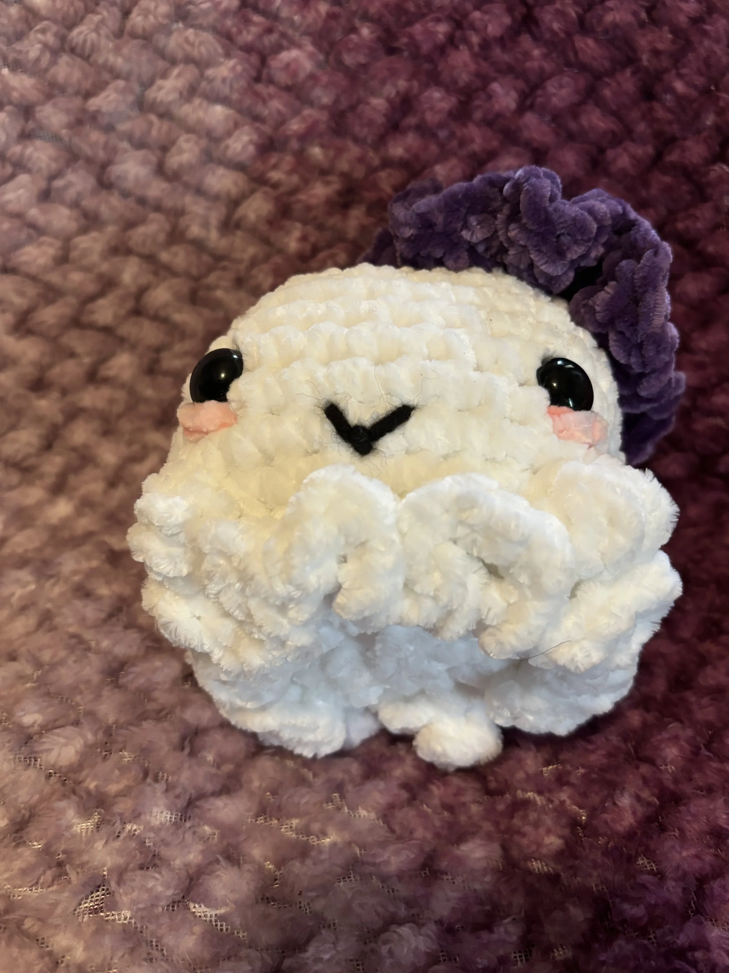 A cute, round plush toy resembling a white fluffy animal with big black eyes, small pink cheeks, and a tiny smiling mouth, wearing a purple beret, on a textured purple blanket.