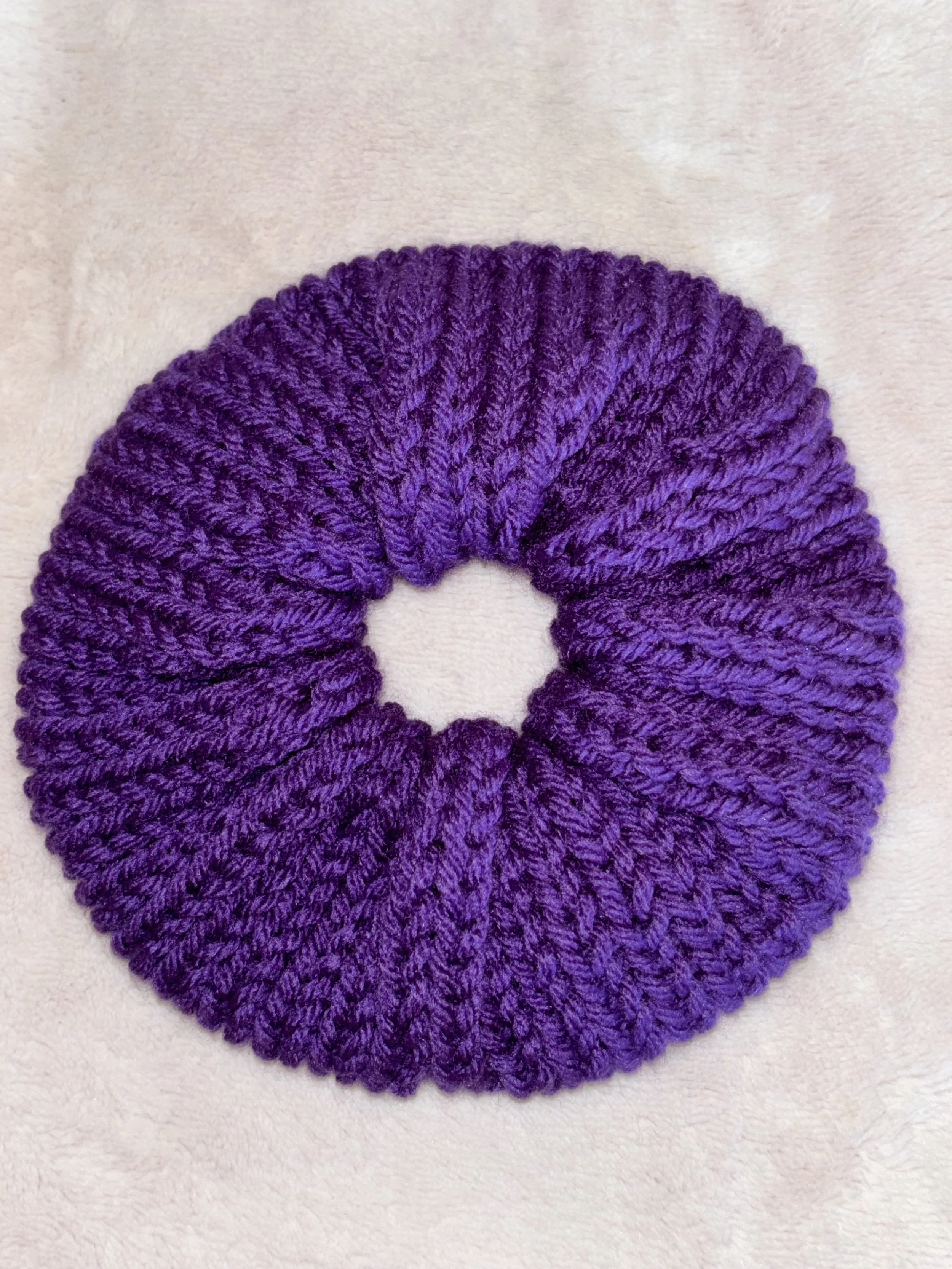 A purple, yarn-made scrunchie placed on a soft, light-colored fabric background.