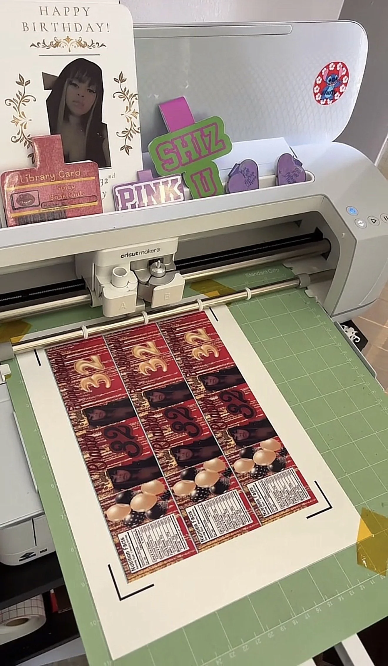 The image shows a Cricut cutting machine preparing a sheet of paper with printed labels for a birthday celebration. There are birthday-themed decorations, including a card with a person's photo, and colorful signs with words like 'PINK U' and 'SHIZ'.