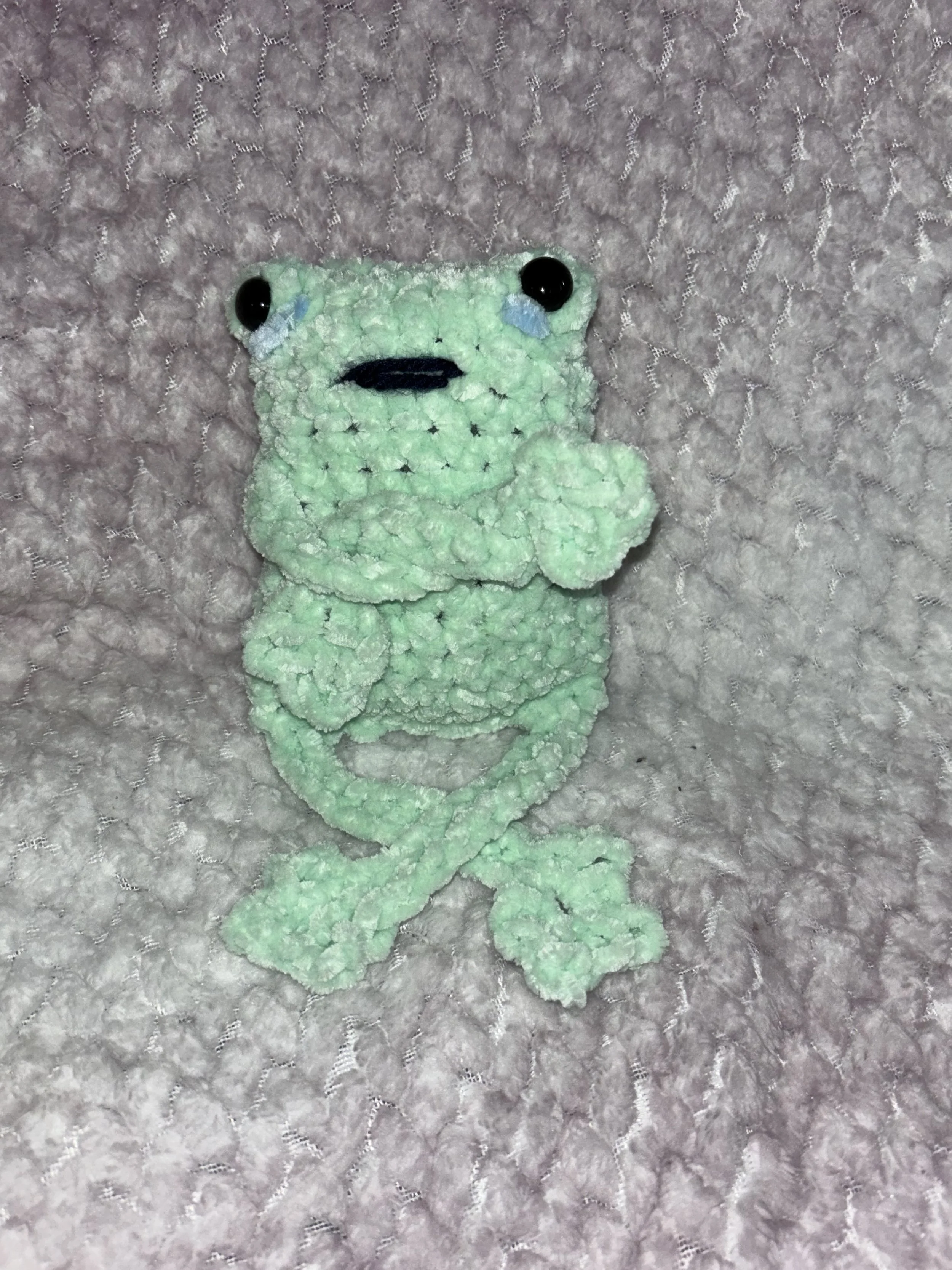 A green, textured frog plush toy with black eyes, a black mouth, and small blue accents near its eyes, resting on a soft, pink, textured fabric background.