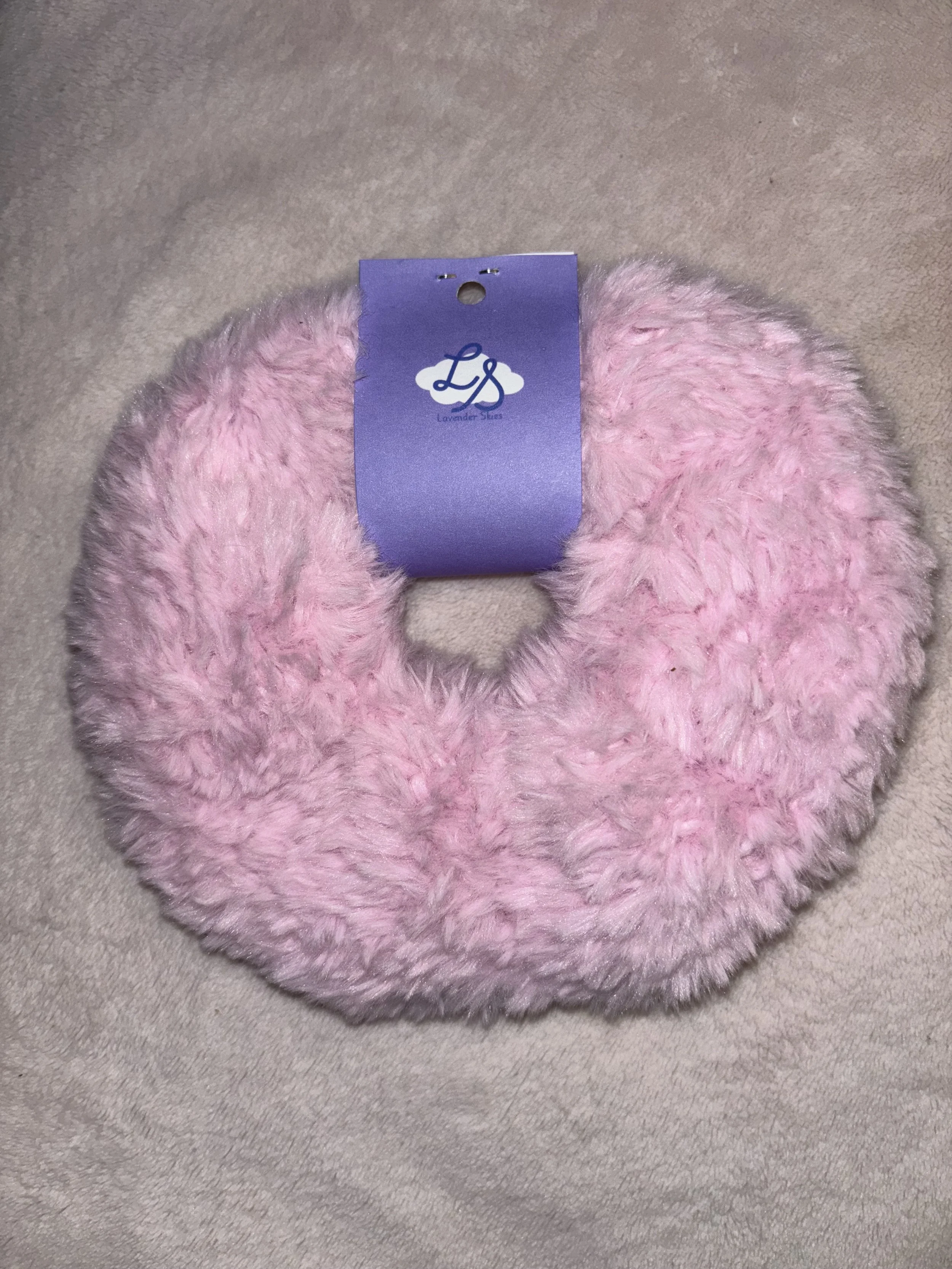 Pink fuzzy scrunchie with a lavender purple tag labeled 'Lavender Skies' resting on a beige fabric surface.