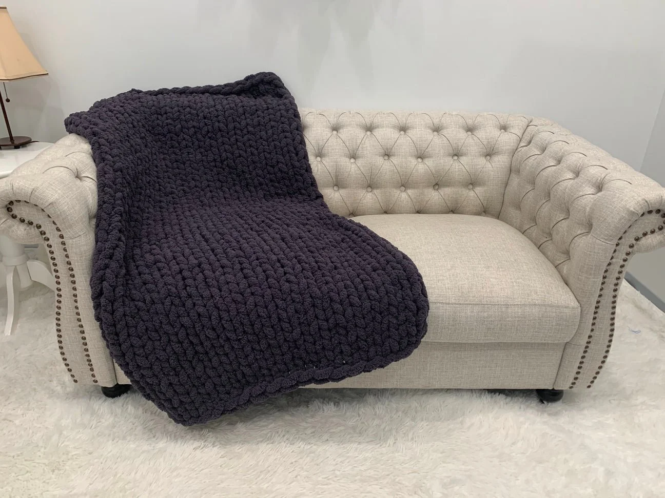 A beige tufted Chesterfield sofa with a dark purple chunky knit blanket draped over the left side, placed on a white shag rug.