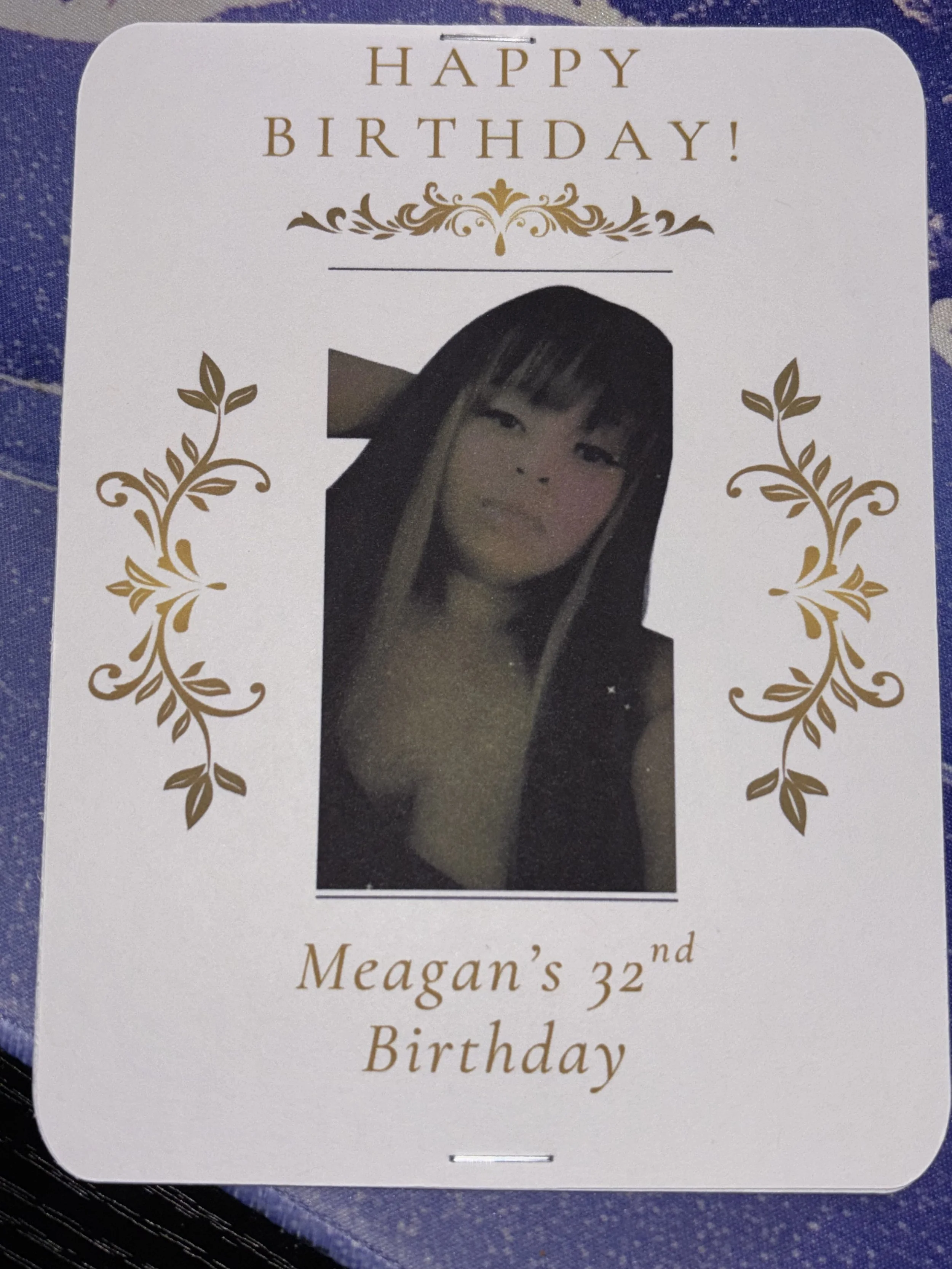 A birthday card with a photo of a young woman with long dark hair and bangs, wearing a dark top. The card has gold decorative elements and text that says "HAPPY BIRTHDAY!" and "Meagan's 32nd Birthday."