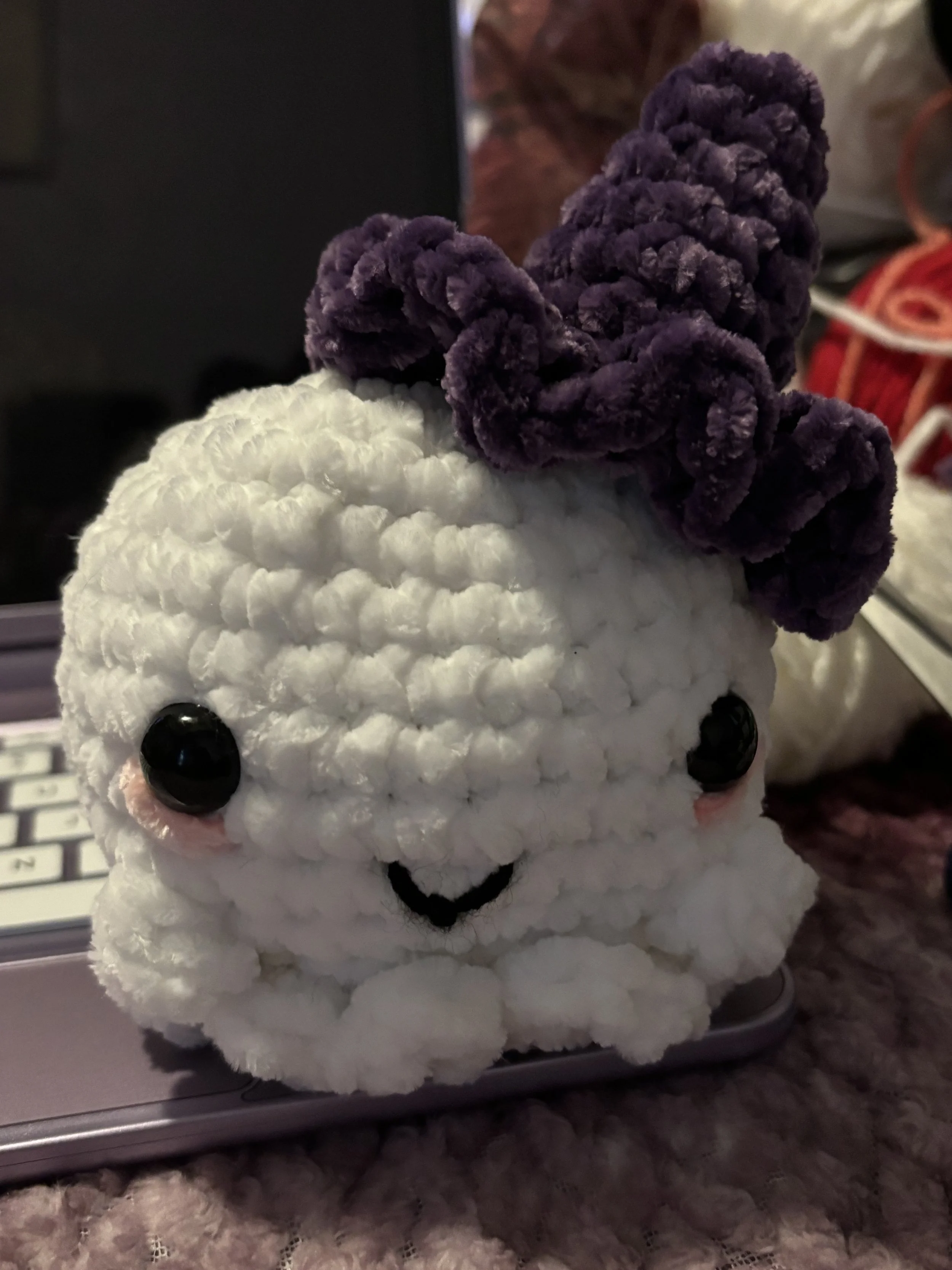 A plush white crocheted bunny with black button eyes, a small embroidered smile, blush patches on the cheeks, and a purple cord bow on its head, placed on a laptop keyboard.