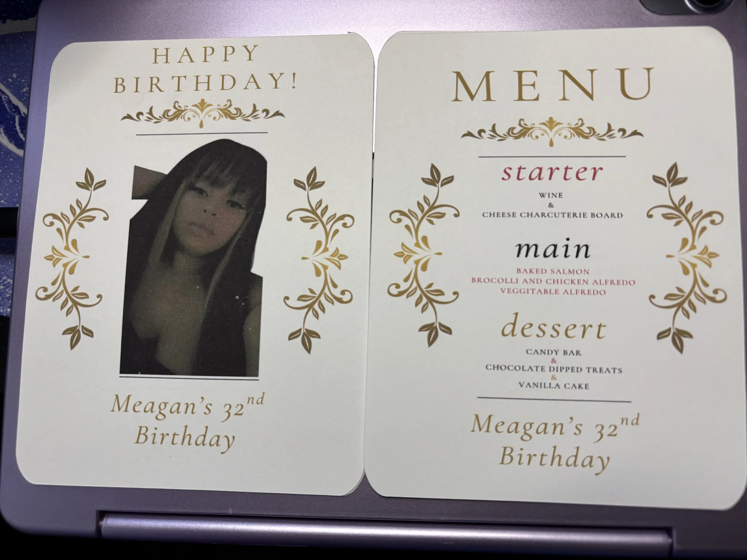 A birthday menu card for Meagan's 32nd birthday. The card features a photo of a woman with dark hair and bangs, decorated with gold floral designs. The menu includes a starter of wine and cheese charcuterie board, main dishes of baked salmon, broccol