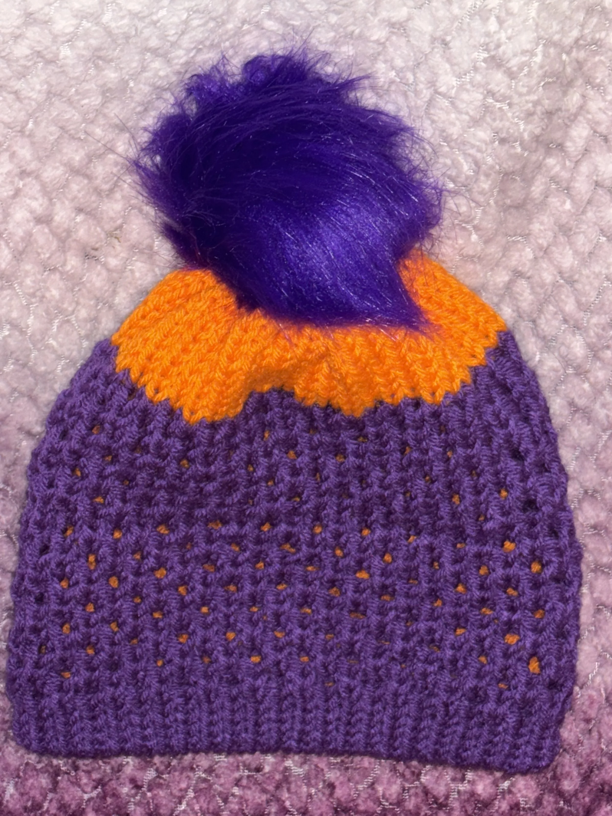 A knitted purple winter hat with orange trim and a purple faux fur pom-pom on top, placed on a pink carpet.