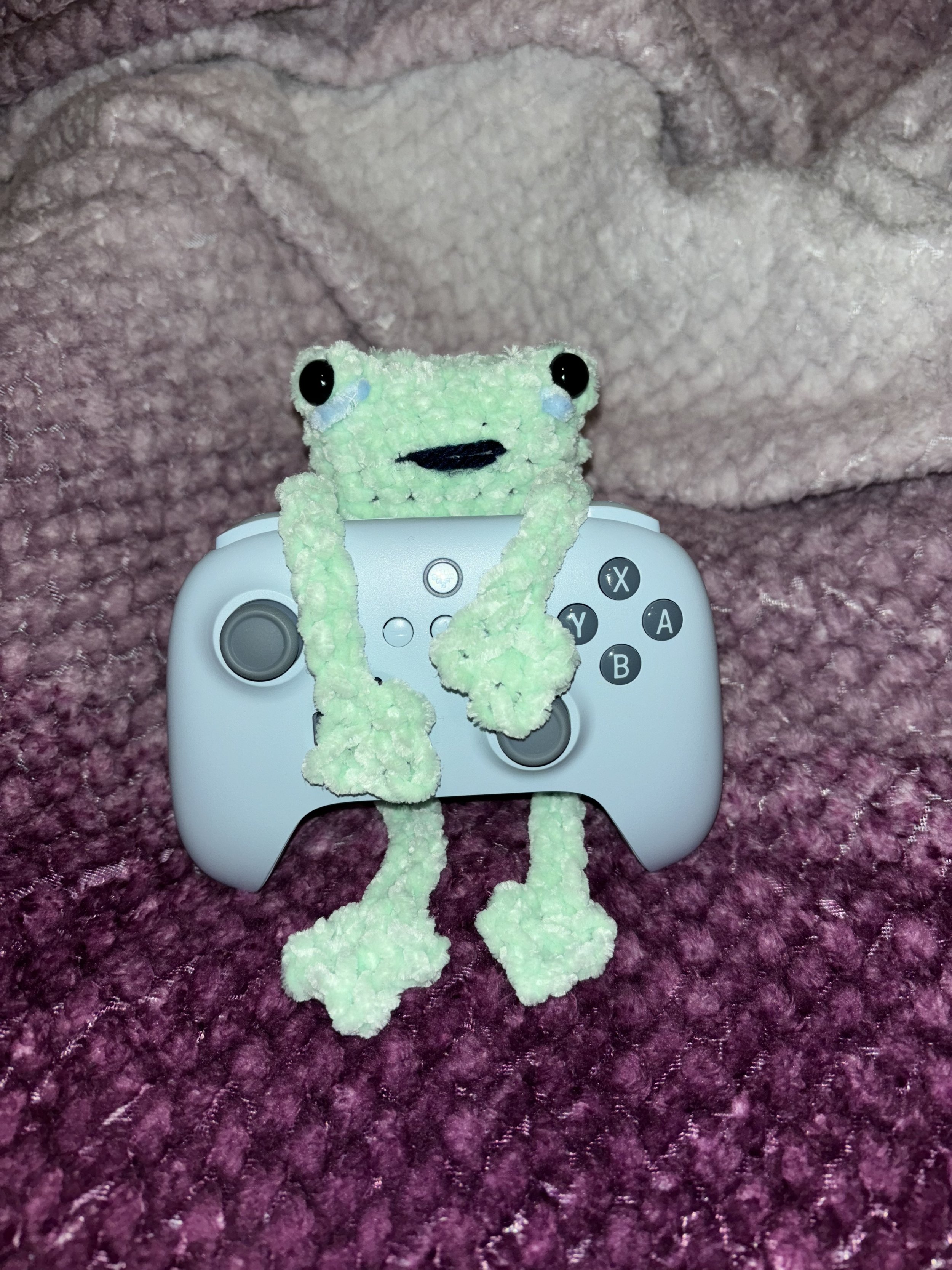 A plush frog holding a gaming controller, sitting on a pink textured surface with a pink fleece blanket in the background.