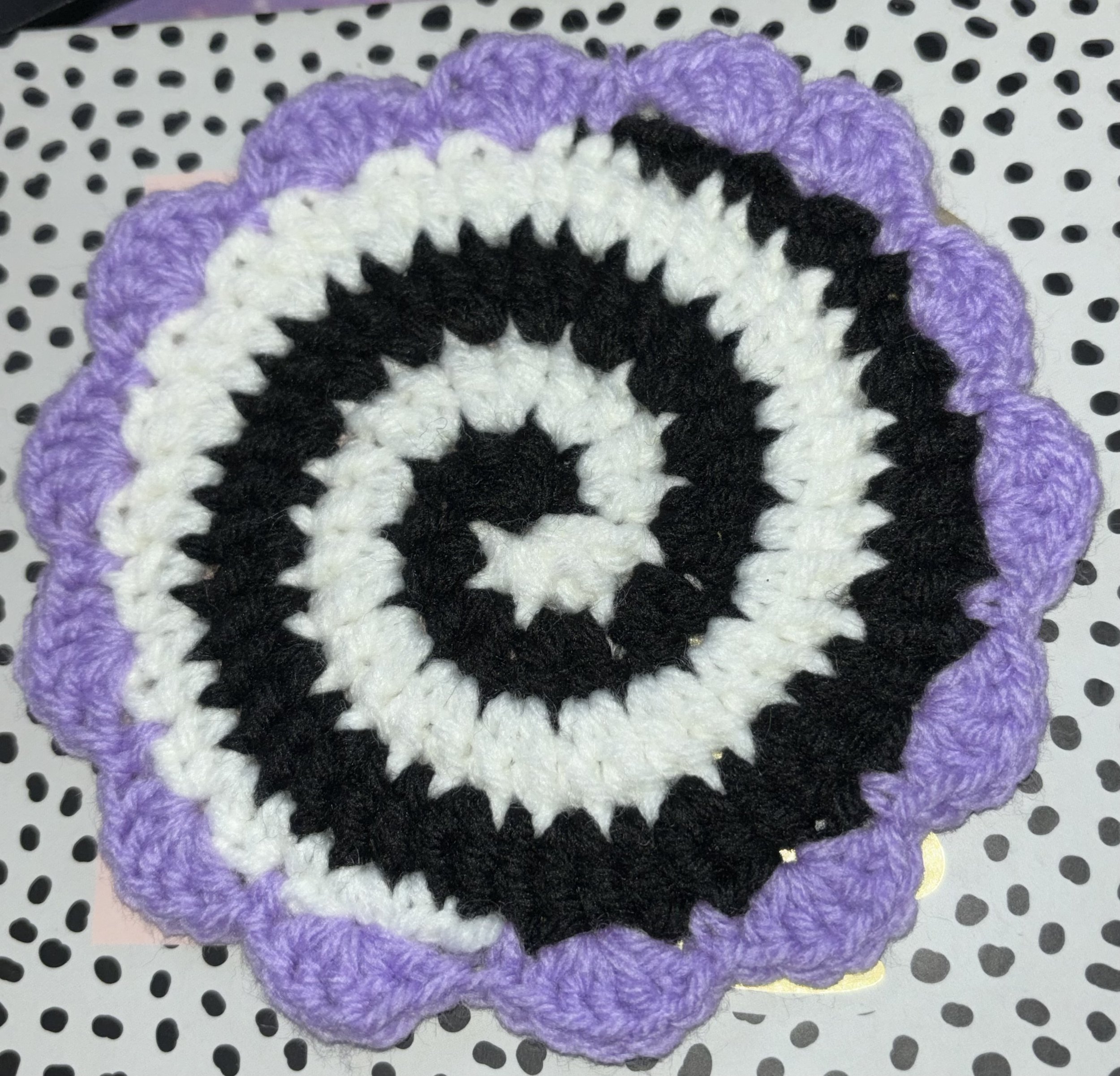 A circular crocheted item with a spiral pattern in black and white yarn, edged with purple yarn, placed on a table with a black and white polka dot surface.