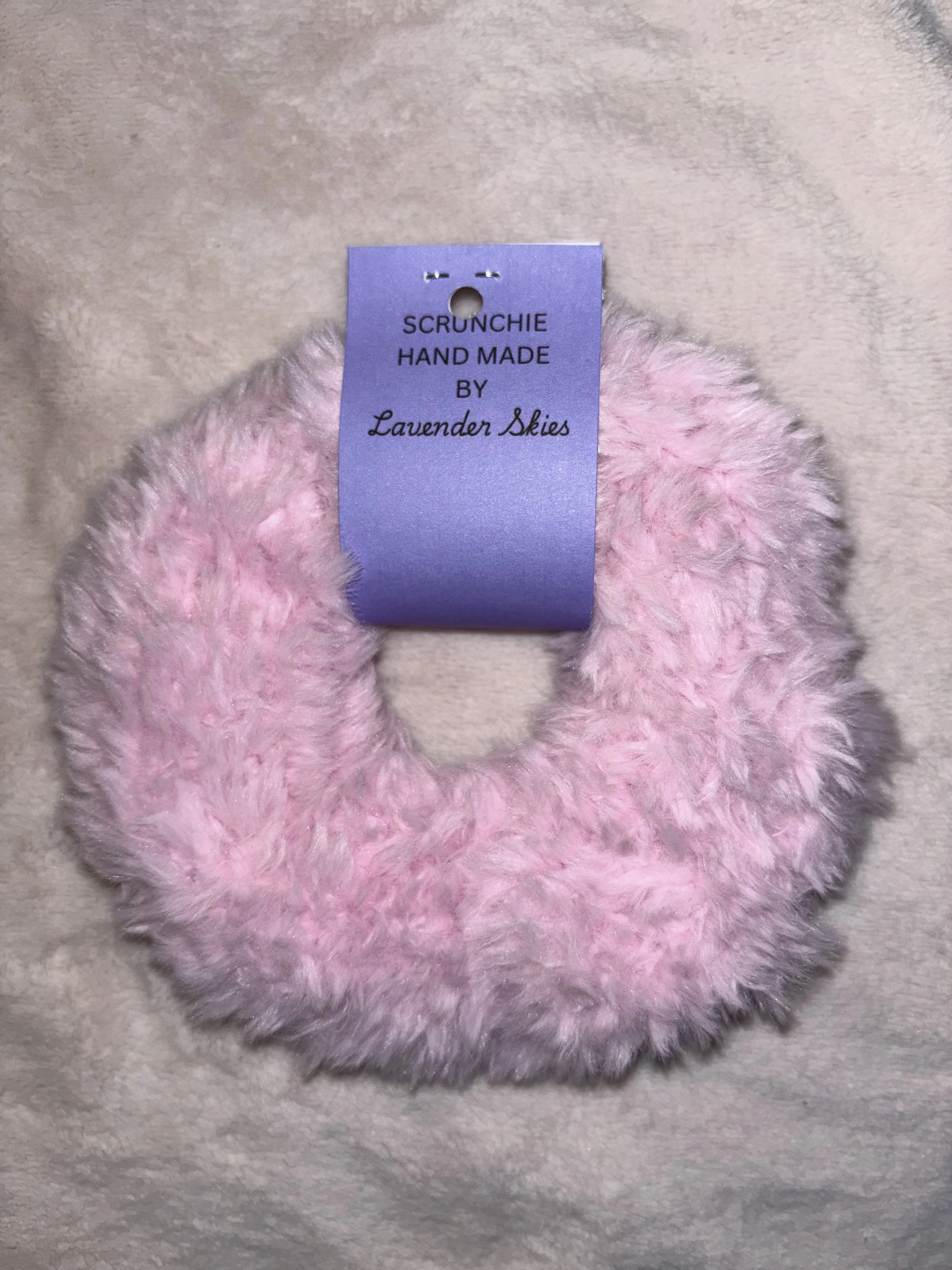A pink, fluffy, circular scrunchie with a purple tag that reads 'SCRUNCHIE HAND MADE BY Lavender Skies'