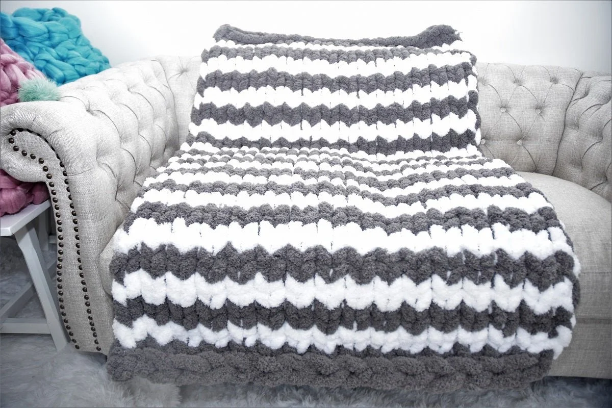 A beige tufted sofa with a black and white striped throw blanket draped over it, and colorful knitted pillows on the armrest, with part of a pink knitted blanket visible on a side table.