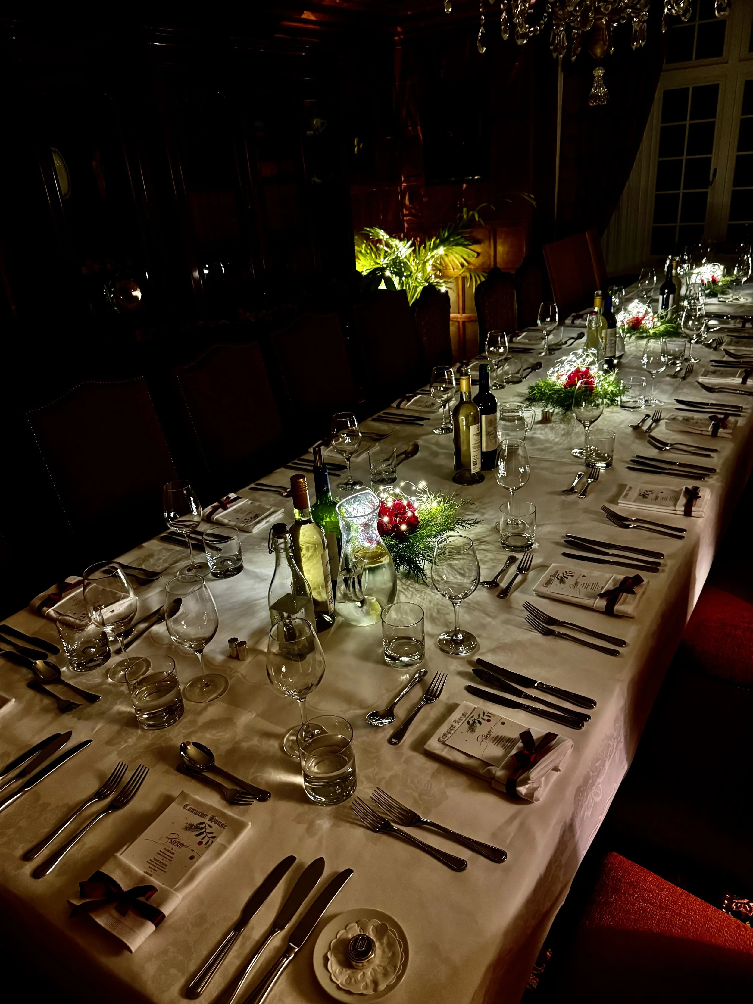 A long dining table set for a formal meal with wine bottles, glasses, silverware, napkins, and floral centerpieces, in a dimly lit room.