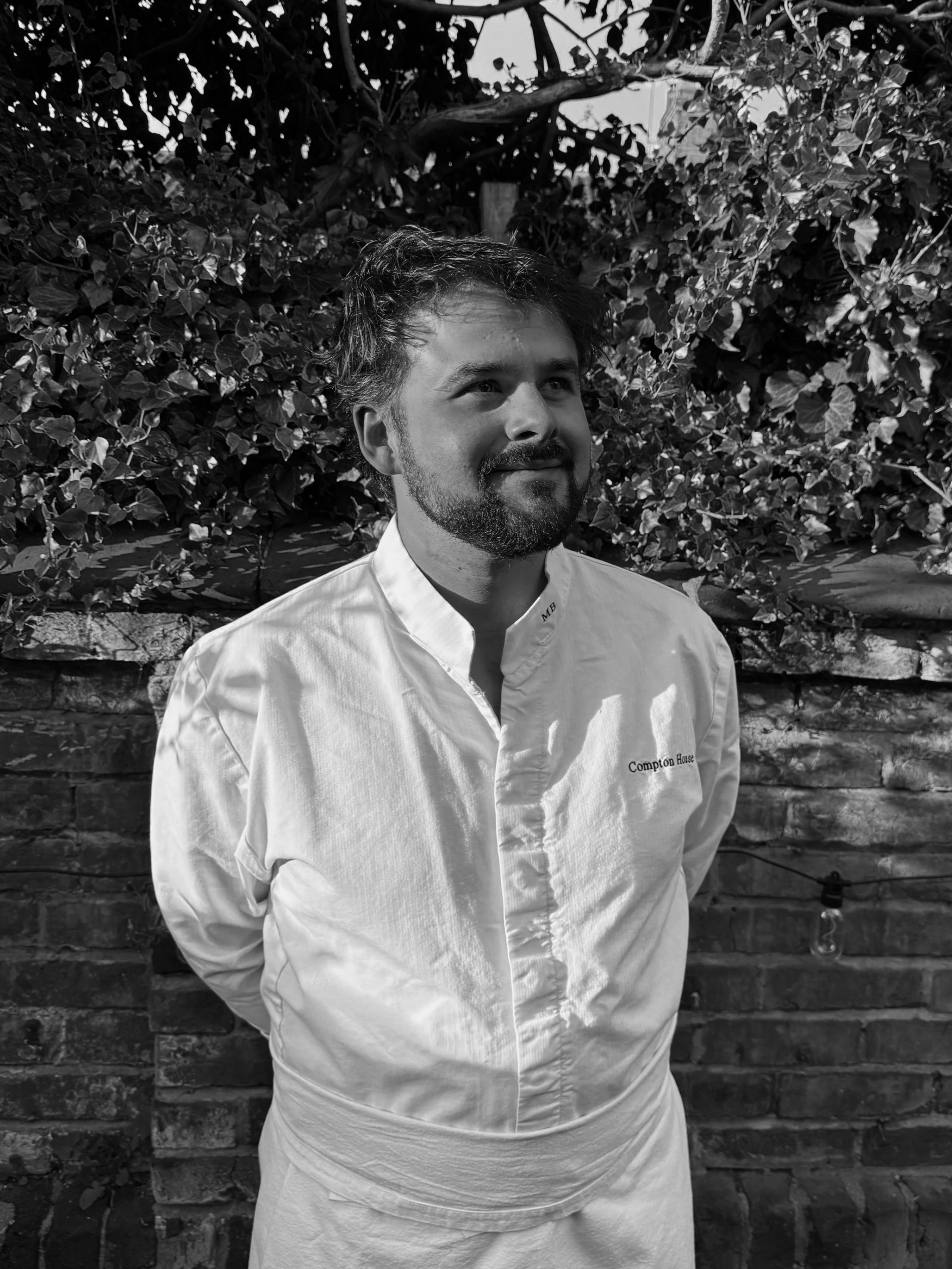 A man with dark, wavy hair and a beard, wearing a white chef's jacket with 'Compton House' embroidered on it, stands outdoors in front of a brick wall and leafy bushes.
