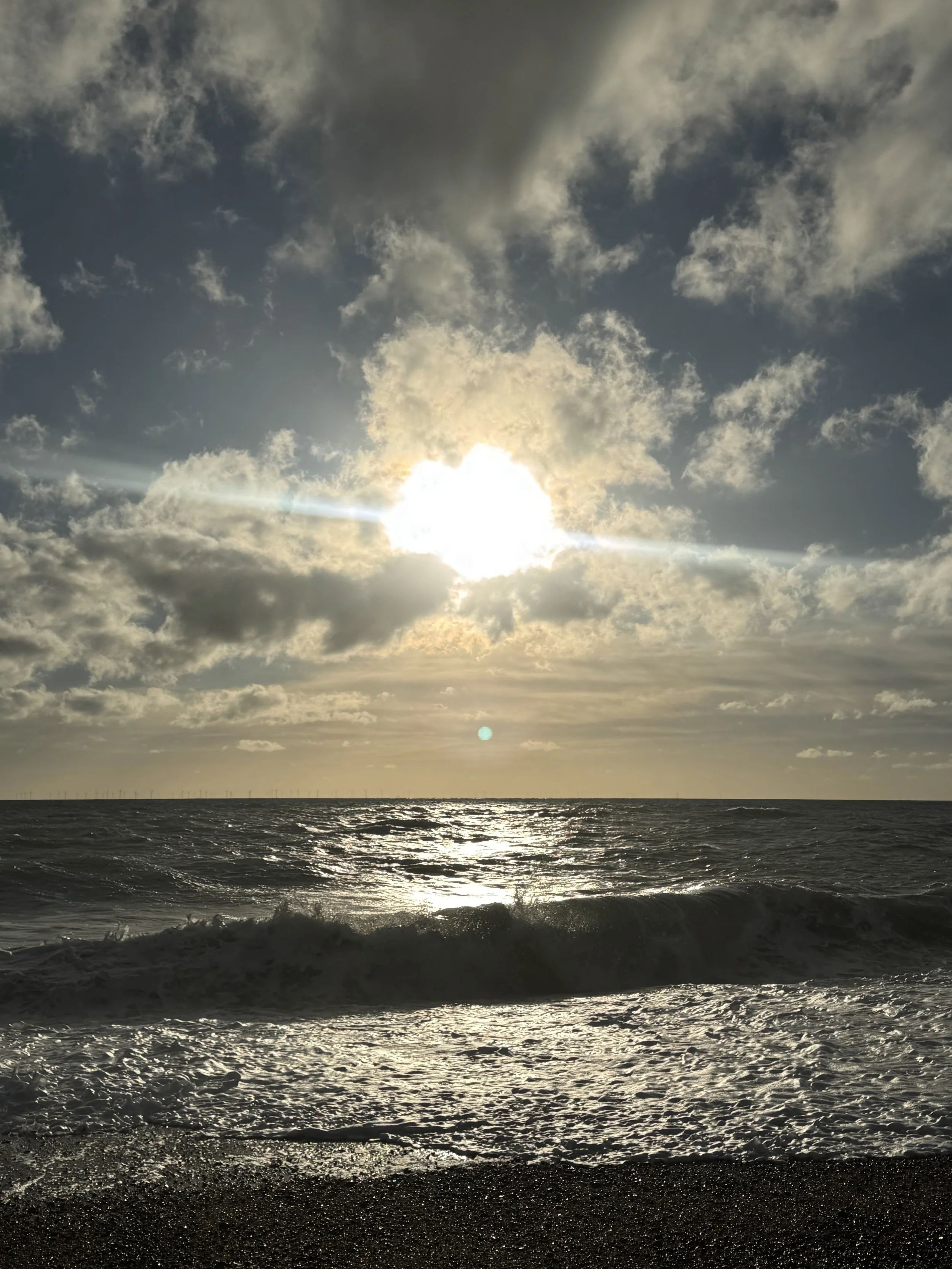 Sun shining through clouds over a rough ocean with waves crashing on the shore.