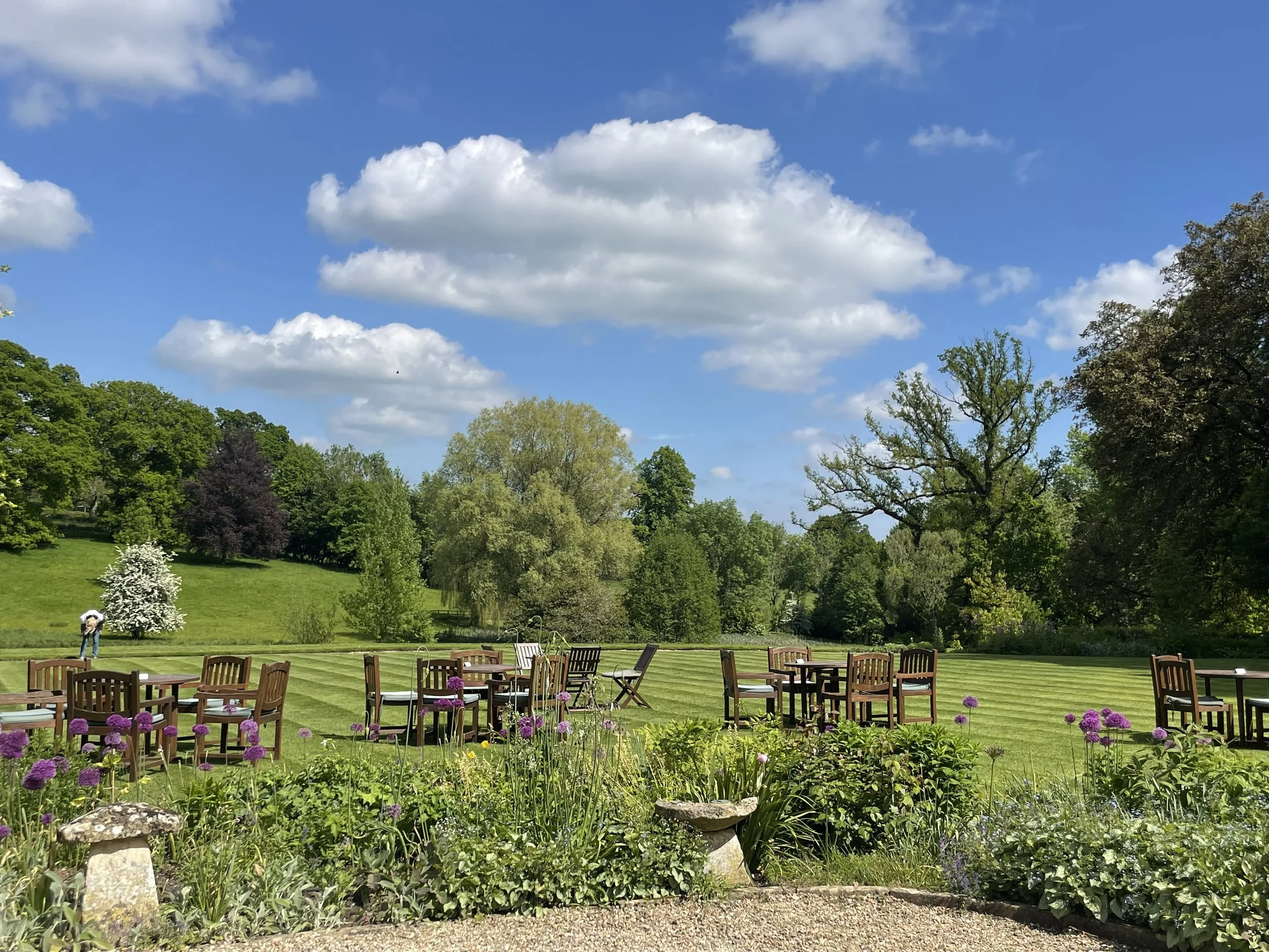A scenic outdoor garden with several wooden tables and chairs on a well-manicured lawn, surrounded by colorful flowers and lush green trees under a partly cloudy blue sky.