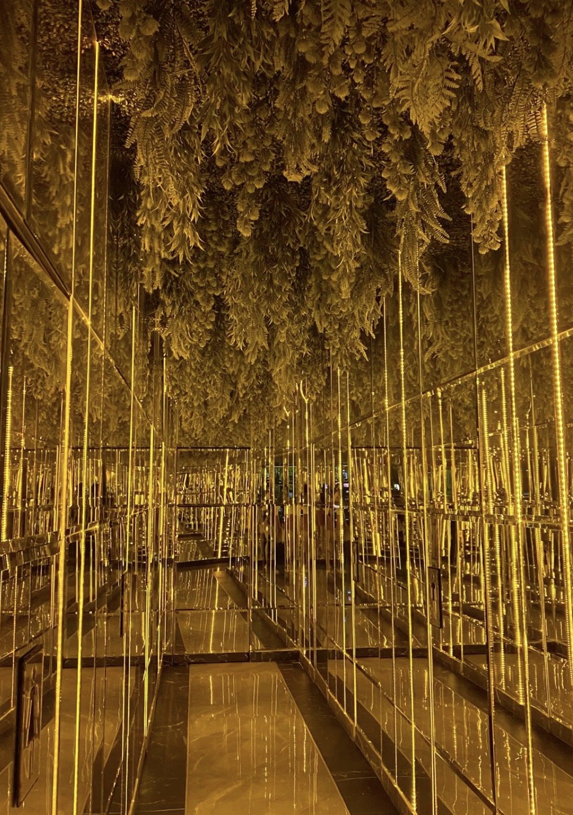 A corridor with mirrored walls and gold-colored walls and ceiling, decorated with artificial greenery and foliage.