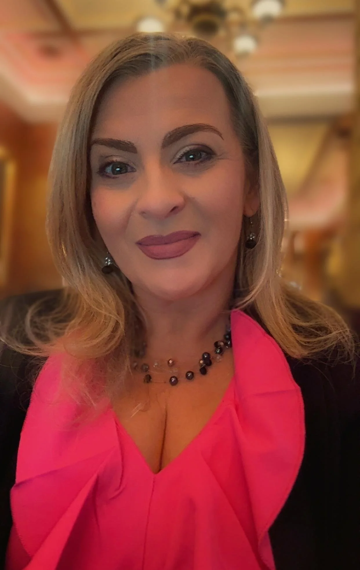 Close-up of a smiling woman with blonde hair, wearing a pink dress, black jewelry, and a black jacket, in a warmly lit indoor setting.