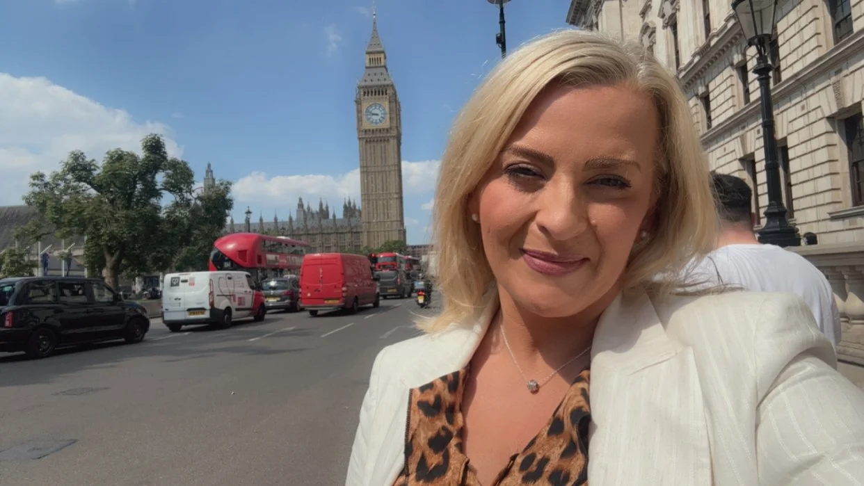 A woman taking a selfie in London with Big Ben and the Houses of Parliament in the background, on a busy street with cars and buses.