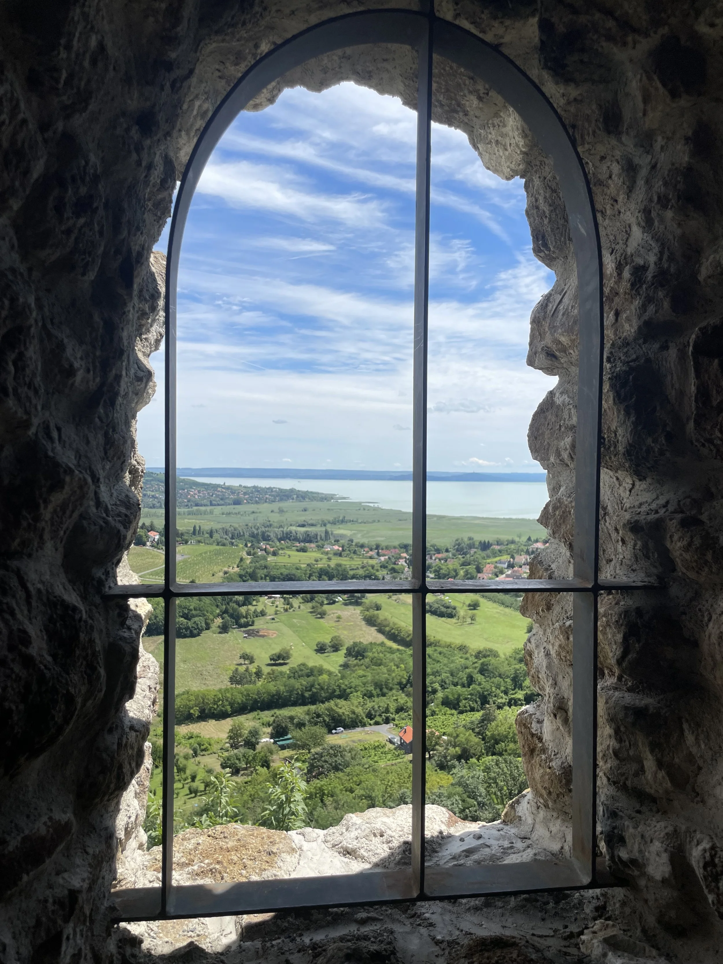 View through a stone window with metal bars showing a landscape of green fields, trees, houses, and a large body of water under a partly cloudy sky.