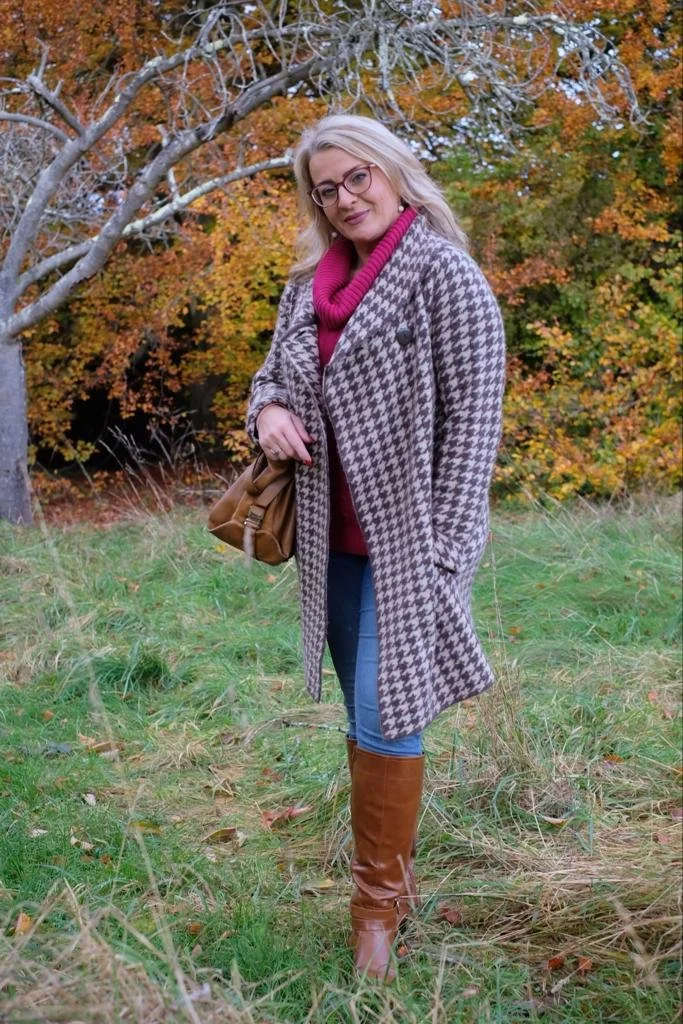A woman with blonde hair, glasses, and wearing a checkered coat over a pink sweater, blue jeans, and brown boots, standing in a grassy area with autumn trees in the background.